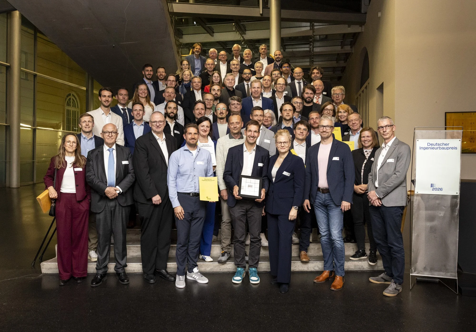 Gruppenfoto aller Preisträgerinnen und Preisträger des Deutschen Ingenieurbaupreises 2026 auf der Treppe im Foyer
