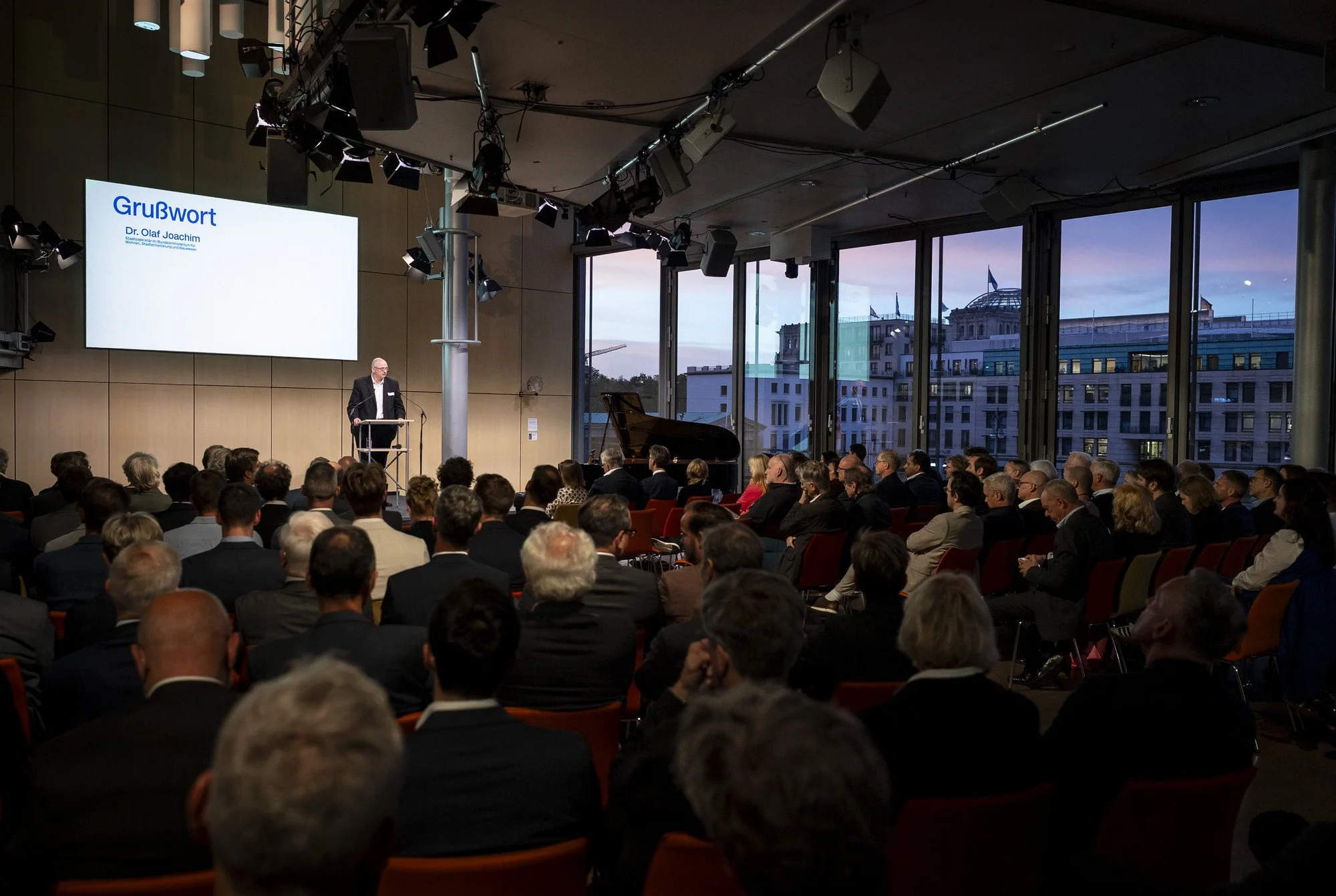 Voll besetzter Plenarsaal der Akademie der Künste in Berlin während der Grußworte, hinter der Glasfront das abendliche Berlin