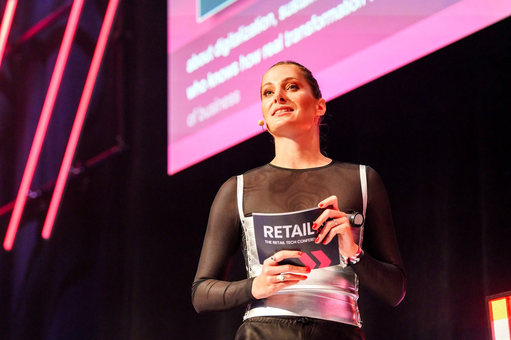 Moderatorin Katharina Aguilar mit Moderationskarte vor pinkfarbener Leinwand auf der Main Stage der Retail-Konferenz RetailNXT 2026