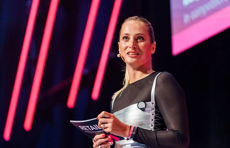 Katharina Aguilar mit Moderationskarte auf der Main Stage der RetailNXT vor pinkfarbener Lichtwand