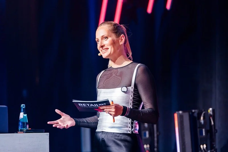Katharina Aguilar spricht mit offener Geste zum Publikum, Moderationskarte der RetailNXT in der Hand