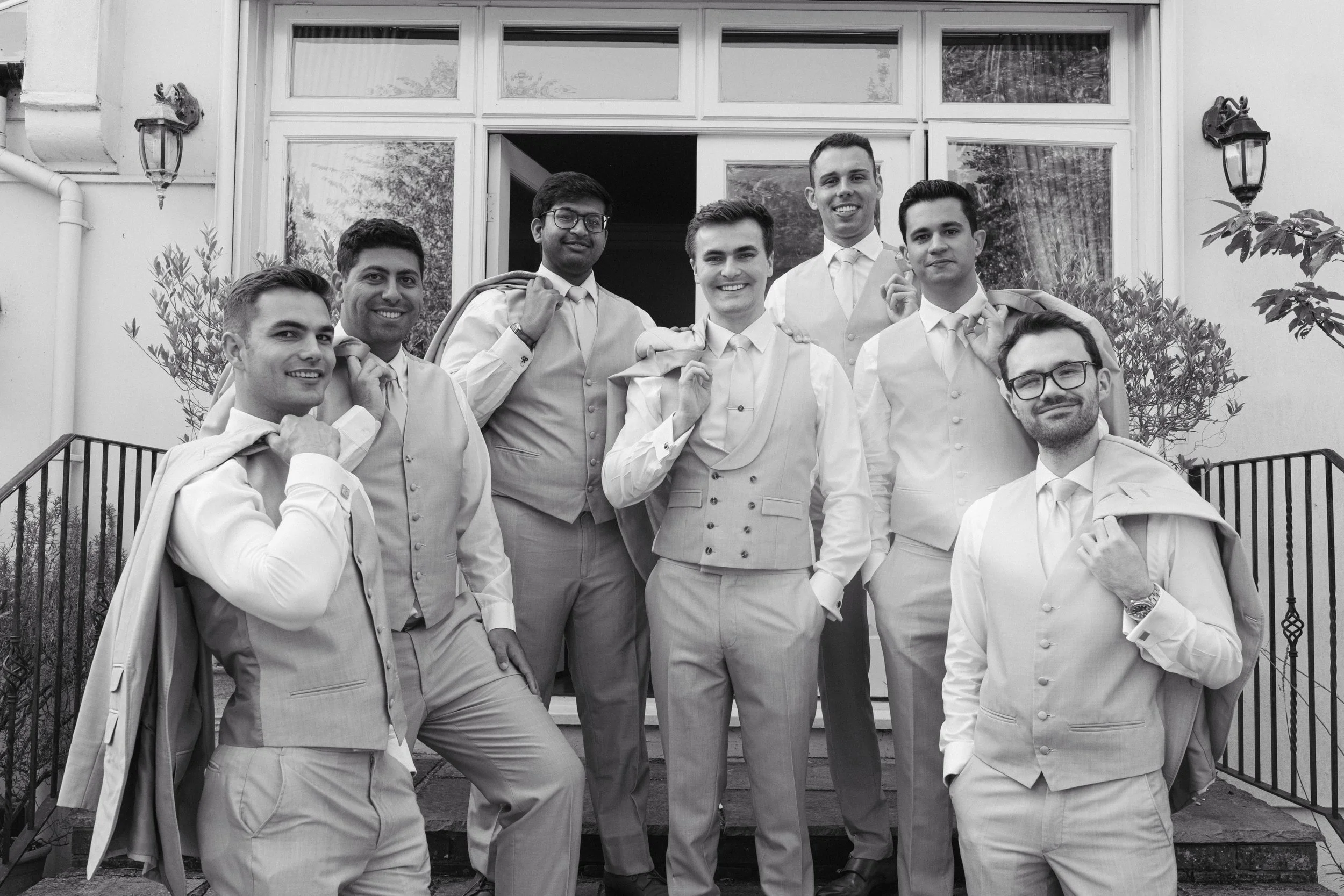 Group of eight men dressed in formal suits, standing on a porch, smiling, some with jackets draped over their shoulders.