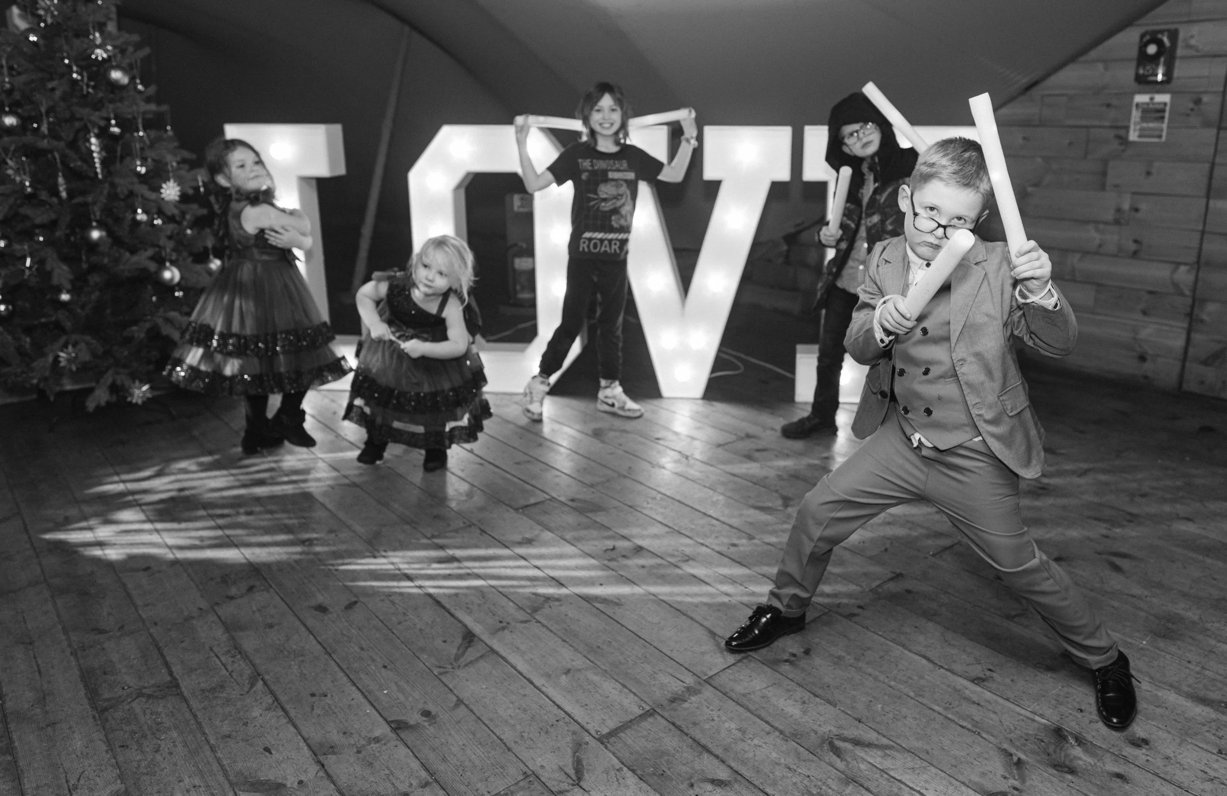 Group of children celebrating Christmas near a decorated tree and large illuminated letters spelling 'LOVE' in the background, with one boy dressed in a suit holding foam sticks in an expressive pose.