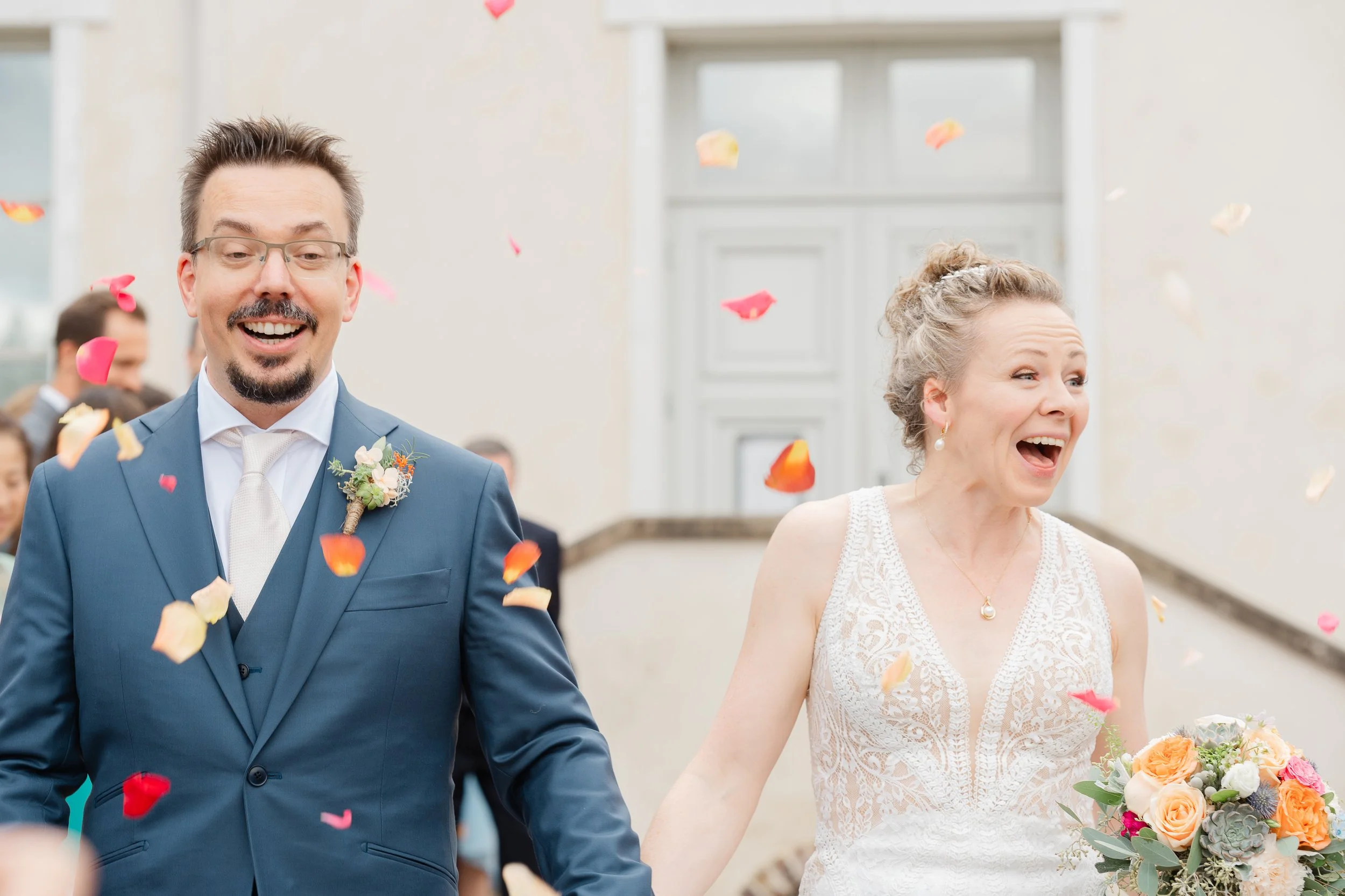 A newly married couple, a man in a blue suit and a woman in a white dress, walking outdoors while throwing rose petals. The man has glasses and a boutonnière, and the woman is holding a bouquet of flowers. They are both smiling with excitement, surro