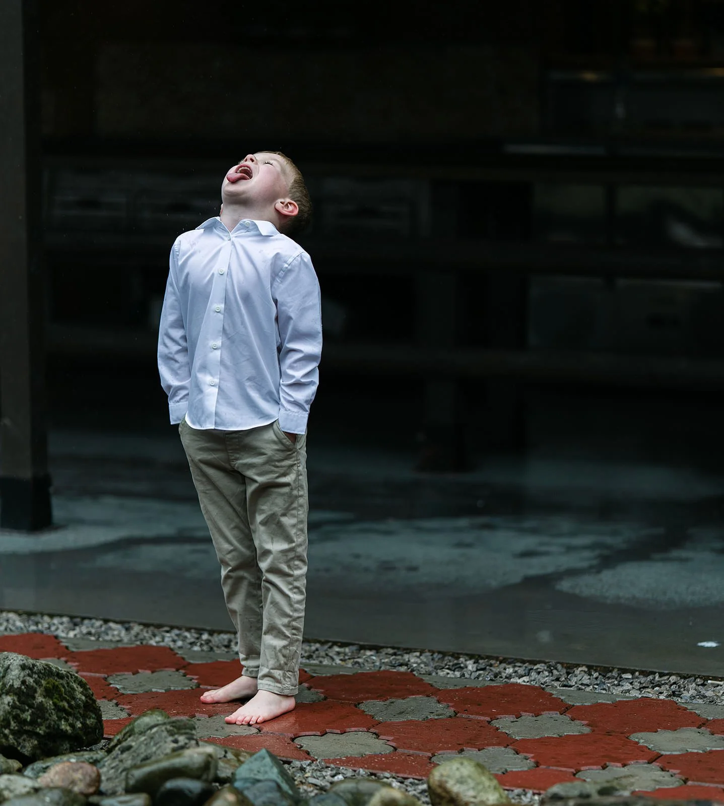 A young boy in a white button-up shirt and beige pants is standing barefoot on a decorative stone pathway, with his head tilted back and tongue sticking out, in an outdoor setting with dark background.