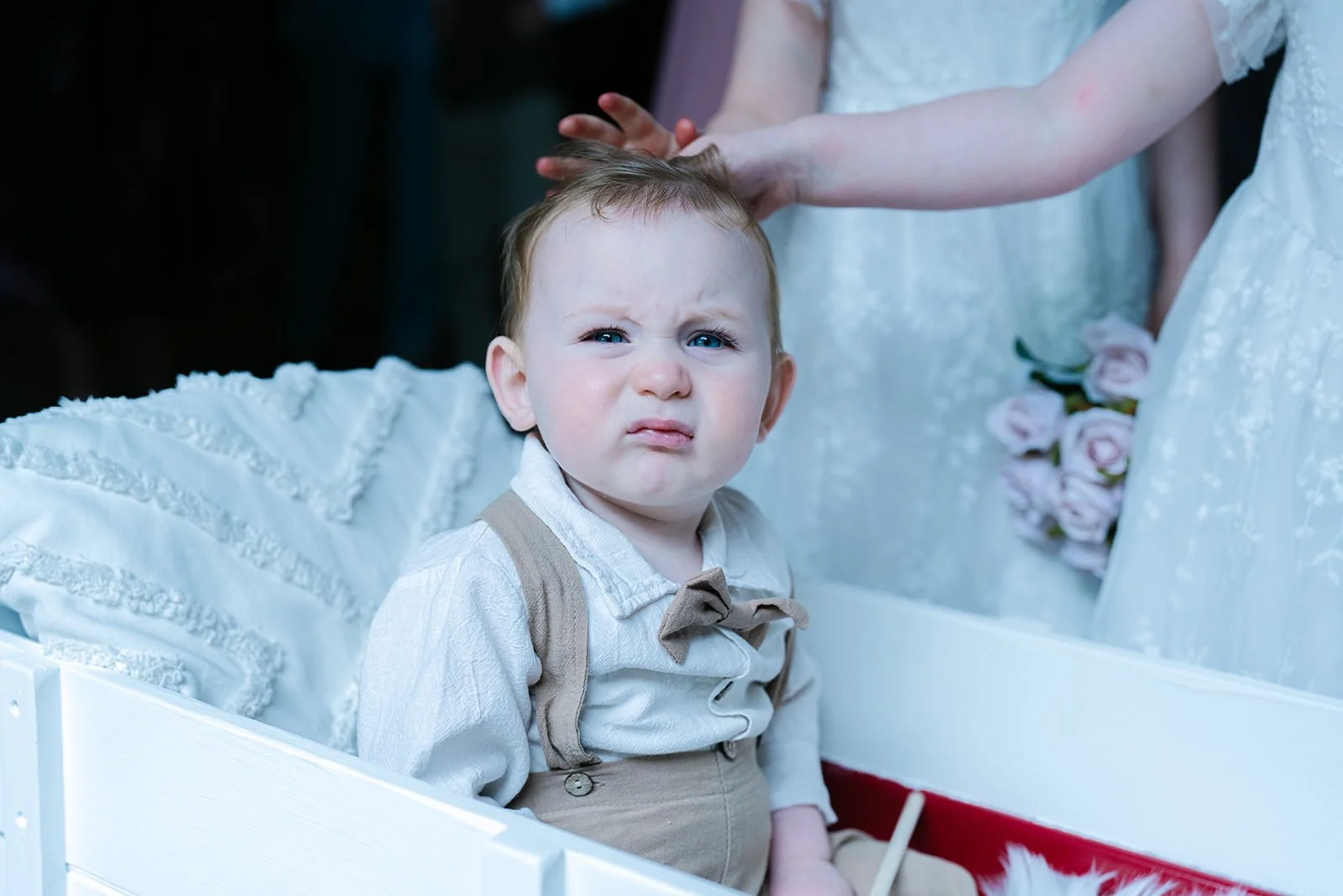 A young boy with a confused or annoyed expression, sitting in a Christmas-themed setting, with people dressed in white and pink roses in the background.