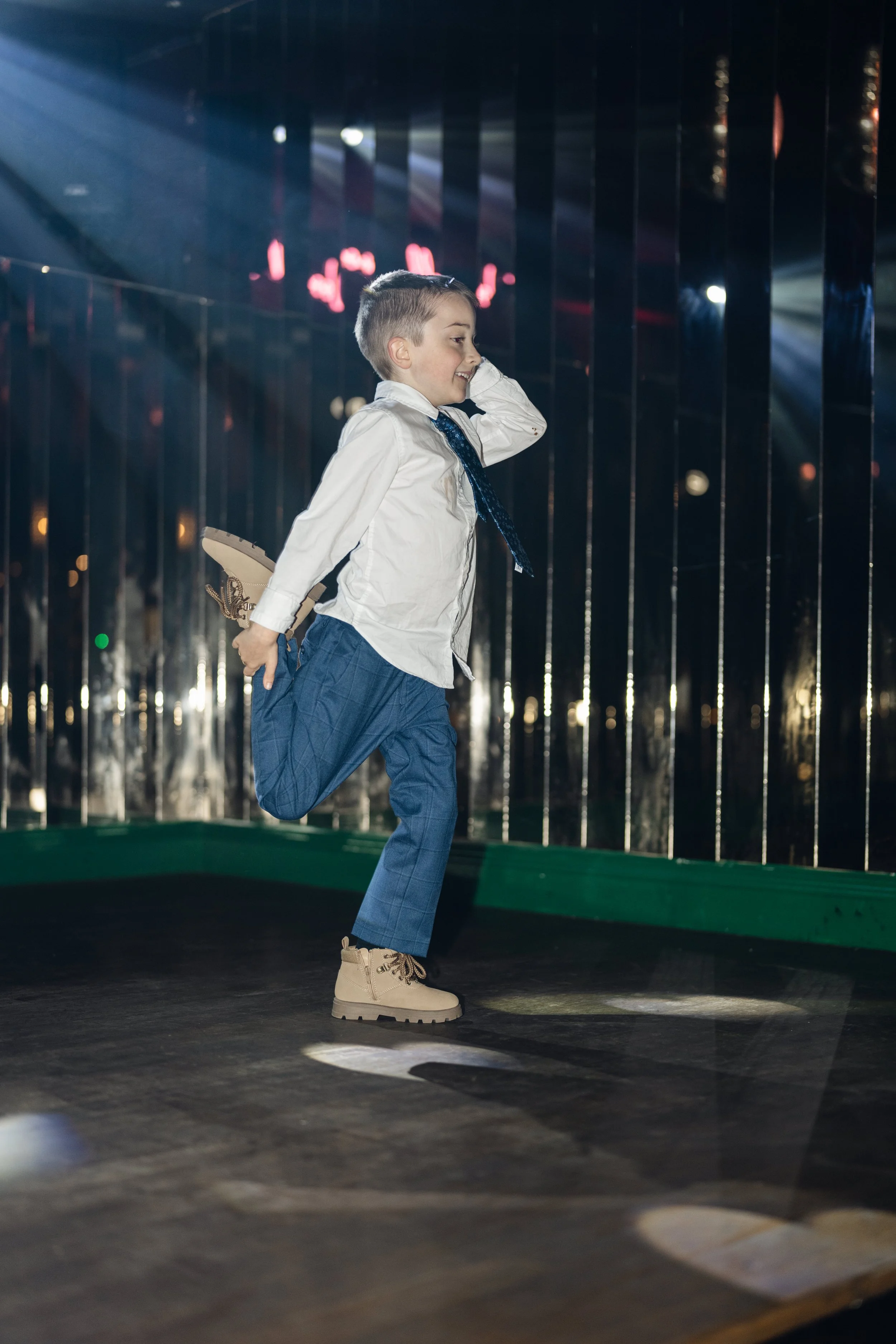 Young boy in dress shirt and blue pants joyfully running on a stage with reflective metallic background and colorful lights.