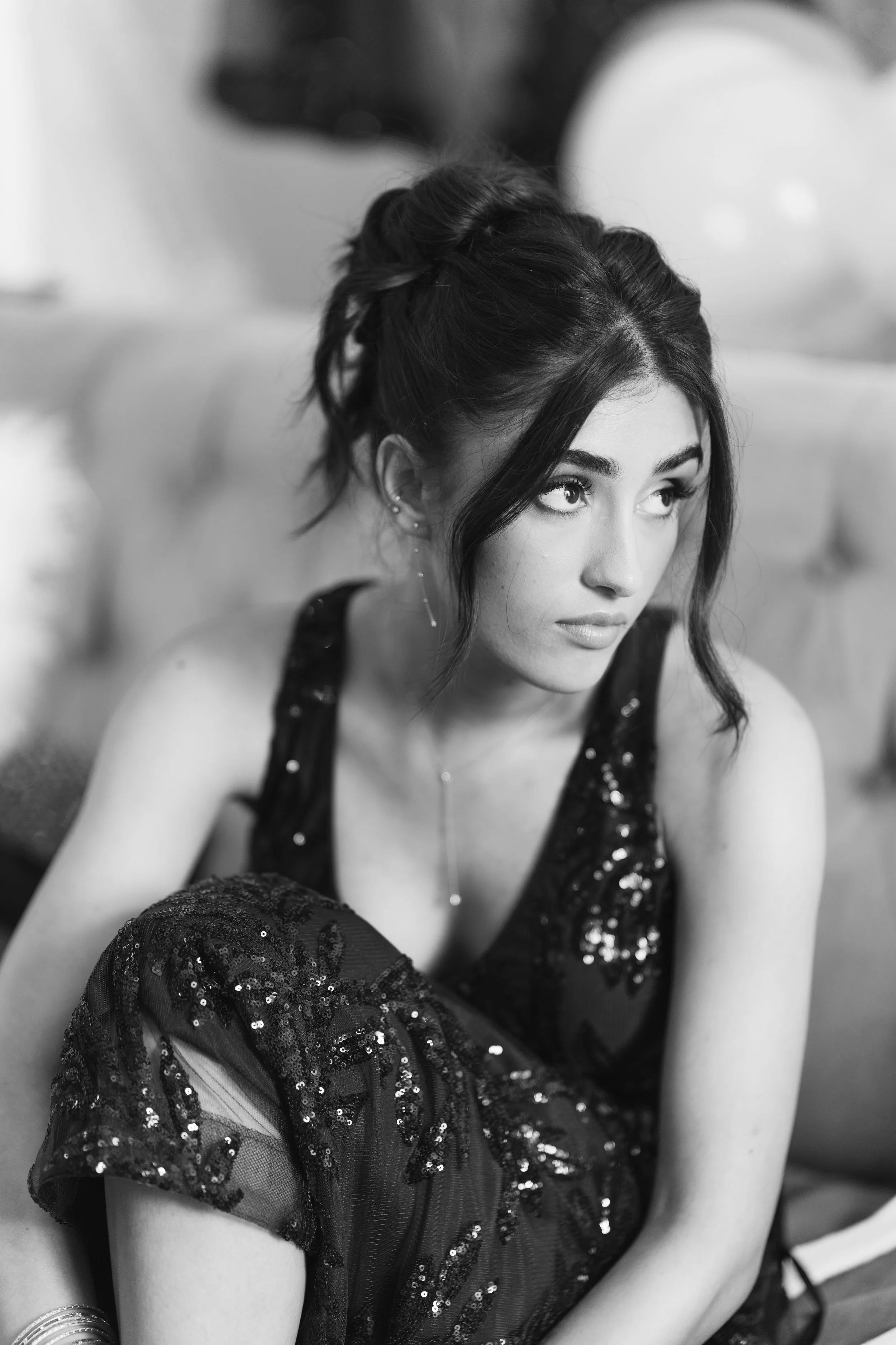 A woman with dark hair styled in a loose updo, wearing a black sequined dress, looking to the side, sitting on a sofa.