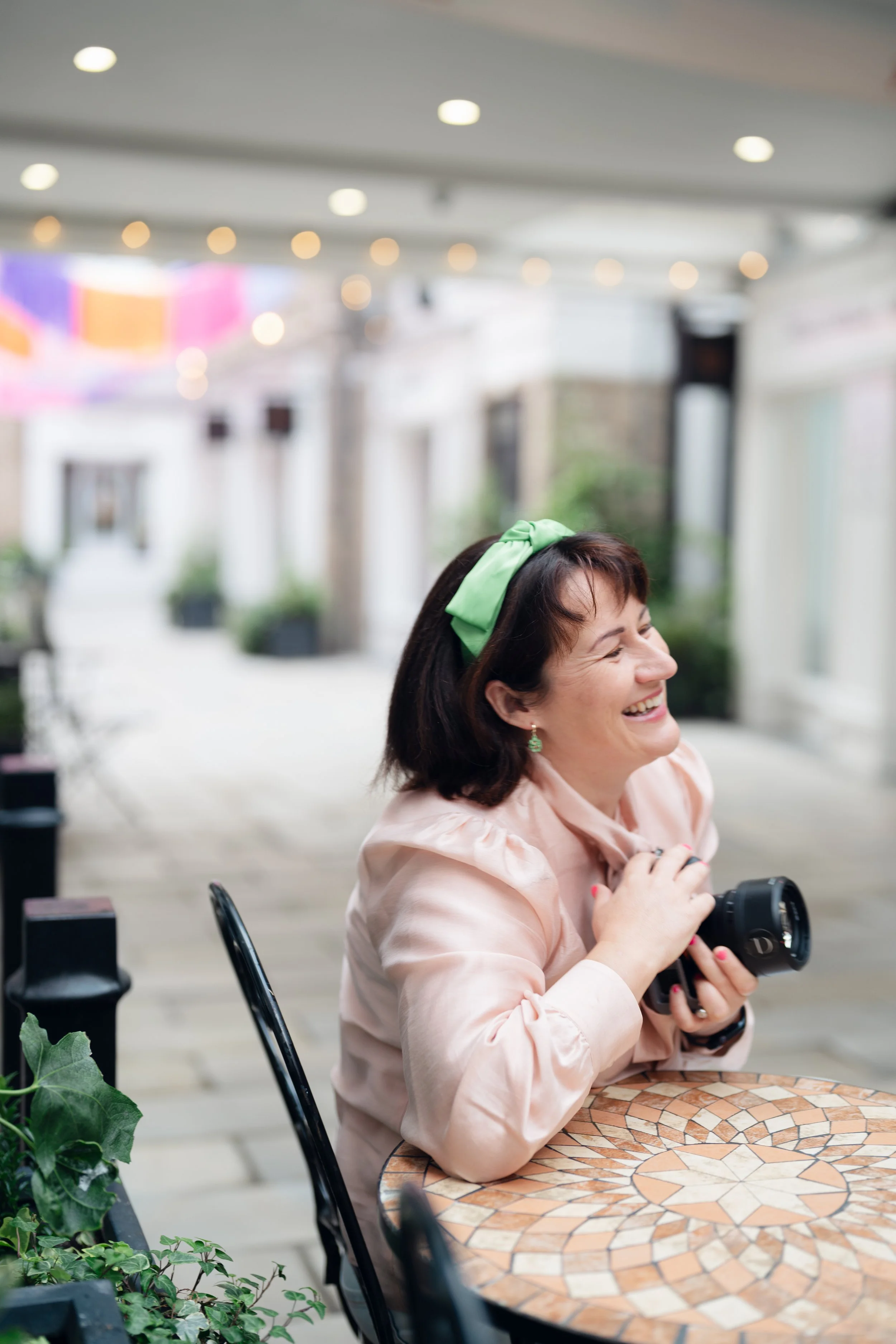 A middle-aged woman with a green headband sitting at a mosaic-top table in a well-lit indoor space, holding a camera and smiling.