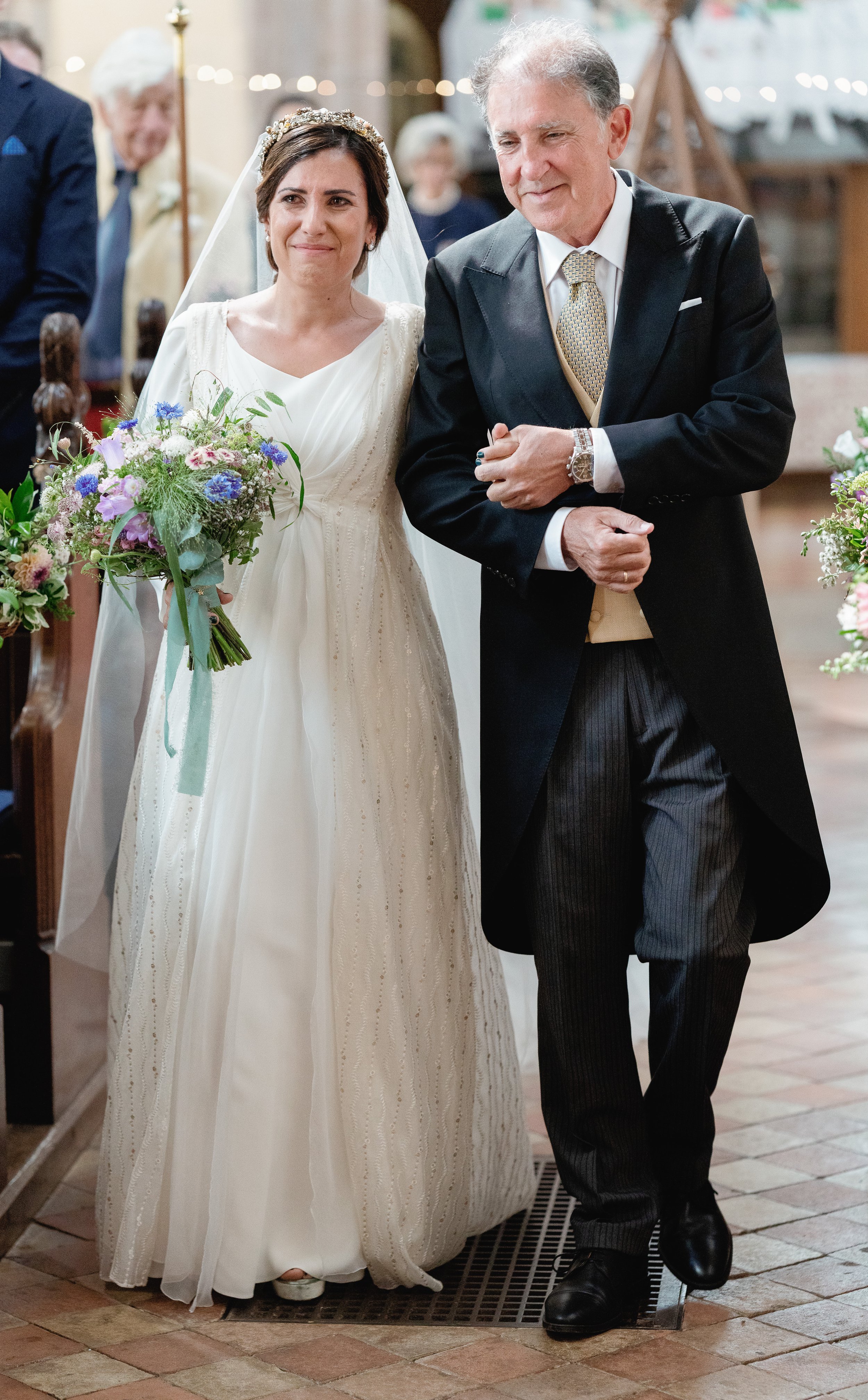 A bride in a white wedding dress holding a bouquet of flowers, walking down the aisle with an older man in a black coat and striped pants during a wedding ceremony in a church.