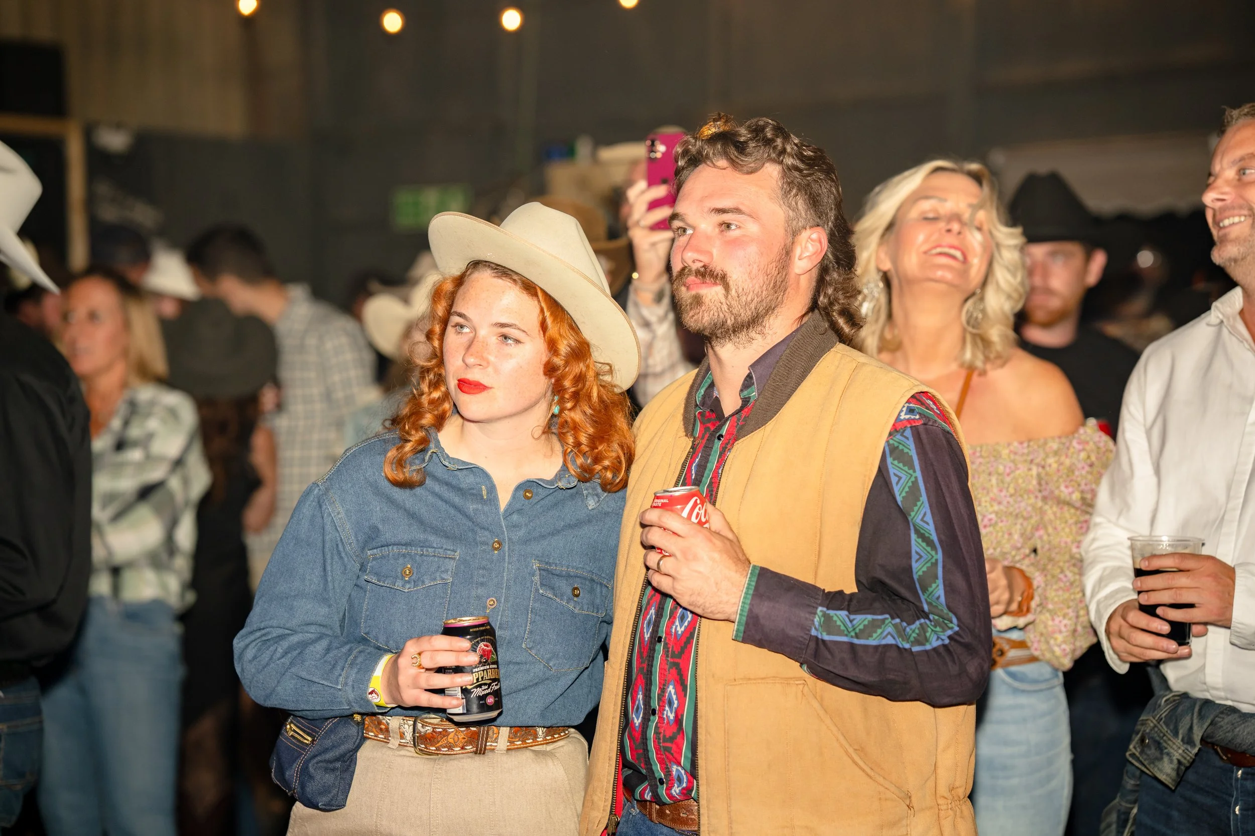 People attending a social event or party, with two individuals in the foreground: a woman with red hair wearing a cowboy hat and denim shirt, and a man with dark hair and beard wearing a colorful vest, holding a soda can and standing close together. 