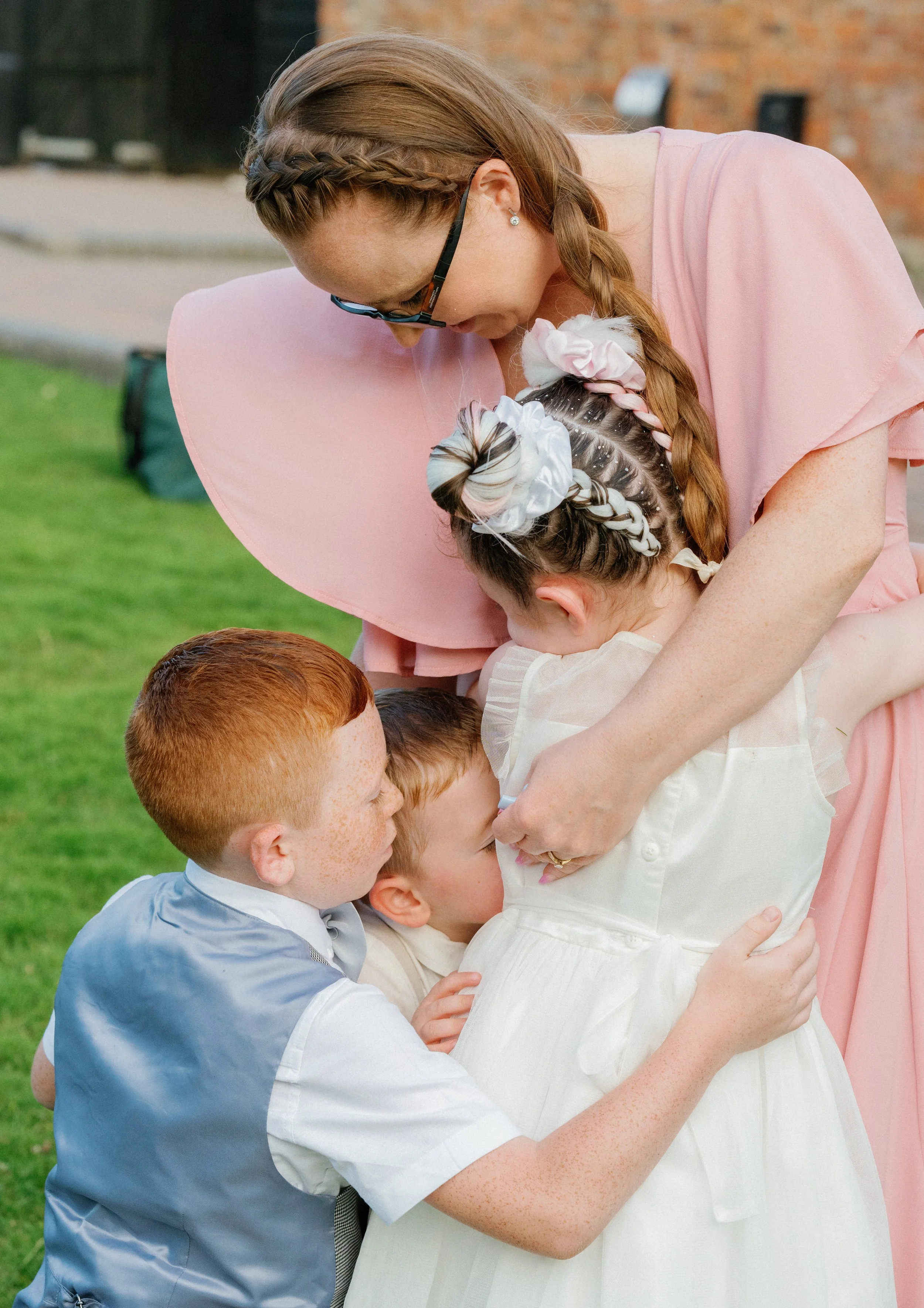 A woman with glasses and a pink dress hugs three children outside on a grassy area, all dressed in formal attire. The children are hugging and embracing in a joyful group hug.
