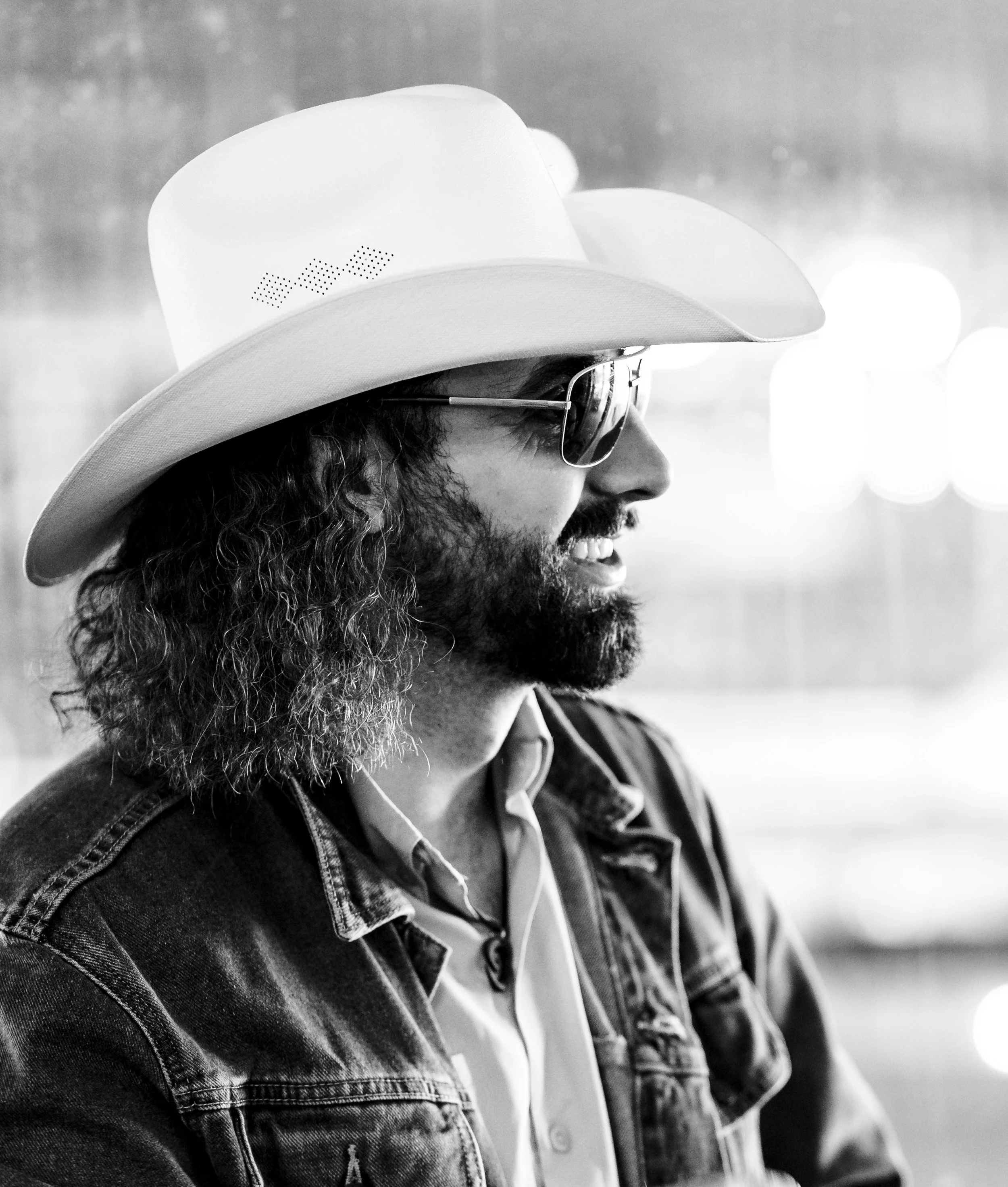 A man with long curly hair, a beard, and glasses smiling while wearing a cowboy hat and denim jacket, with earphones in his ears.