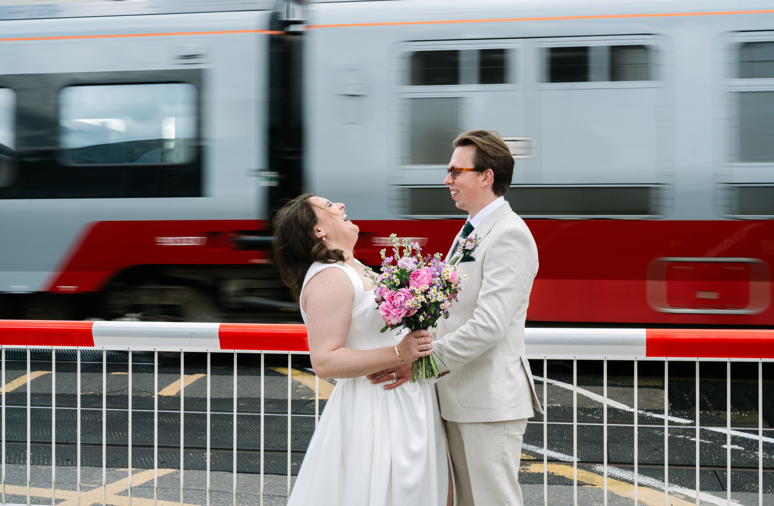 A newlywed couple in wedding attire sharing a joyful moment with a moving train in the background, standing behind a metal barrier.