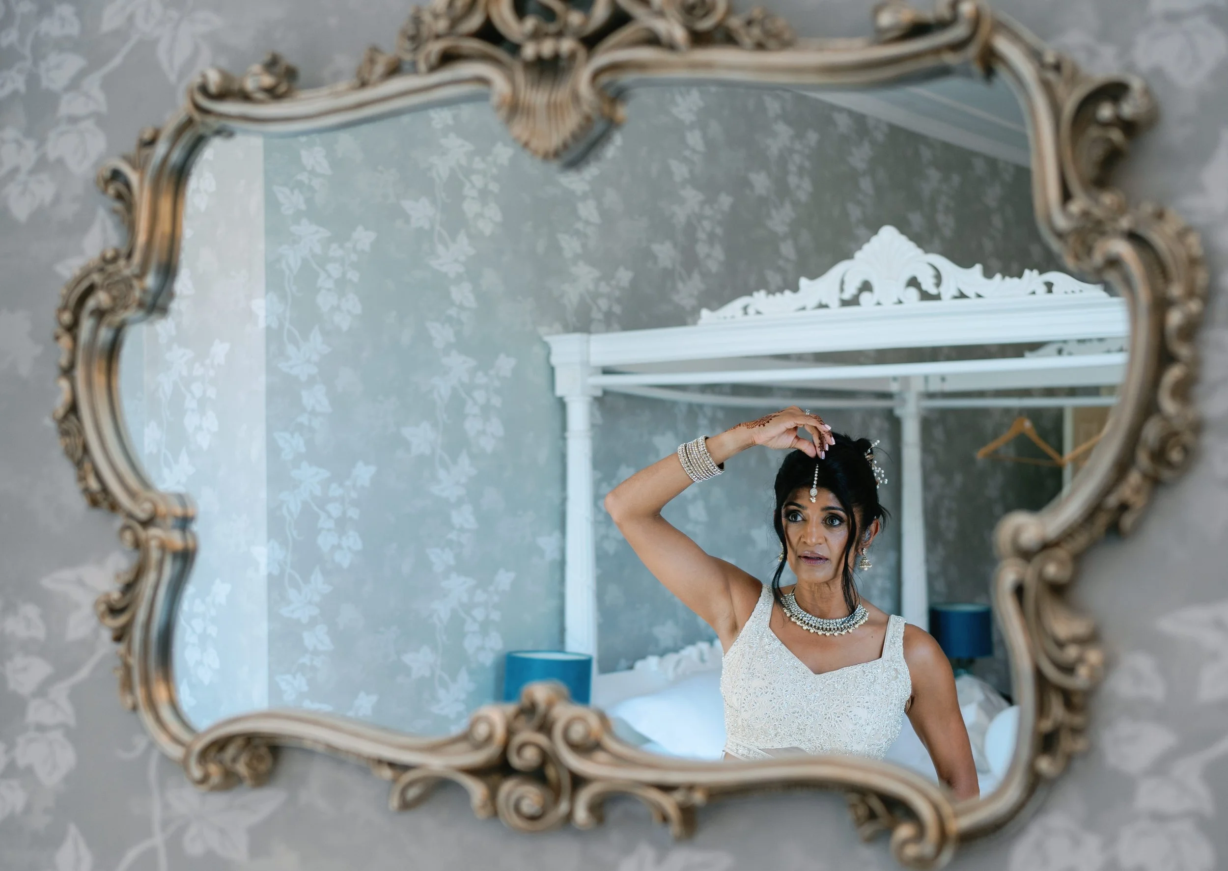 A woman dressed in a white gown and adorned with jewelry is looking at herself in a decorative mirror. She is lifting her hair with one hand while standing in a bedroom with floral wallpaper and a canopy bed in the background.