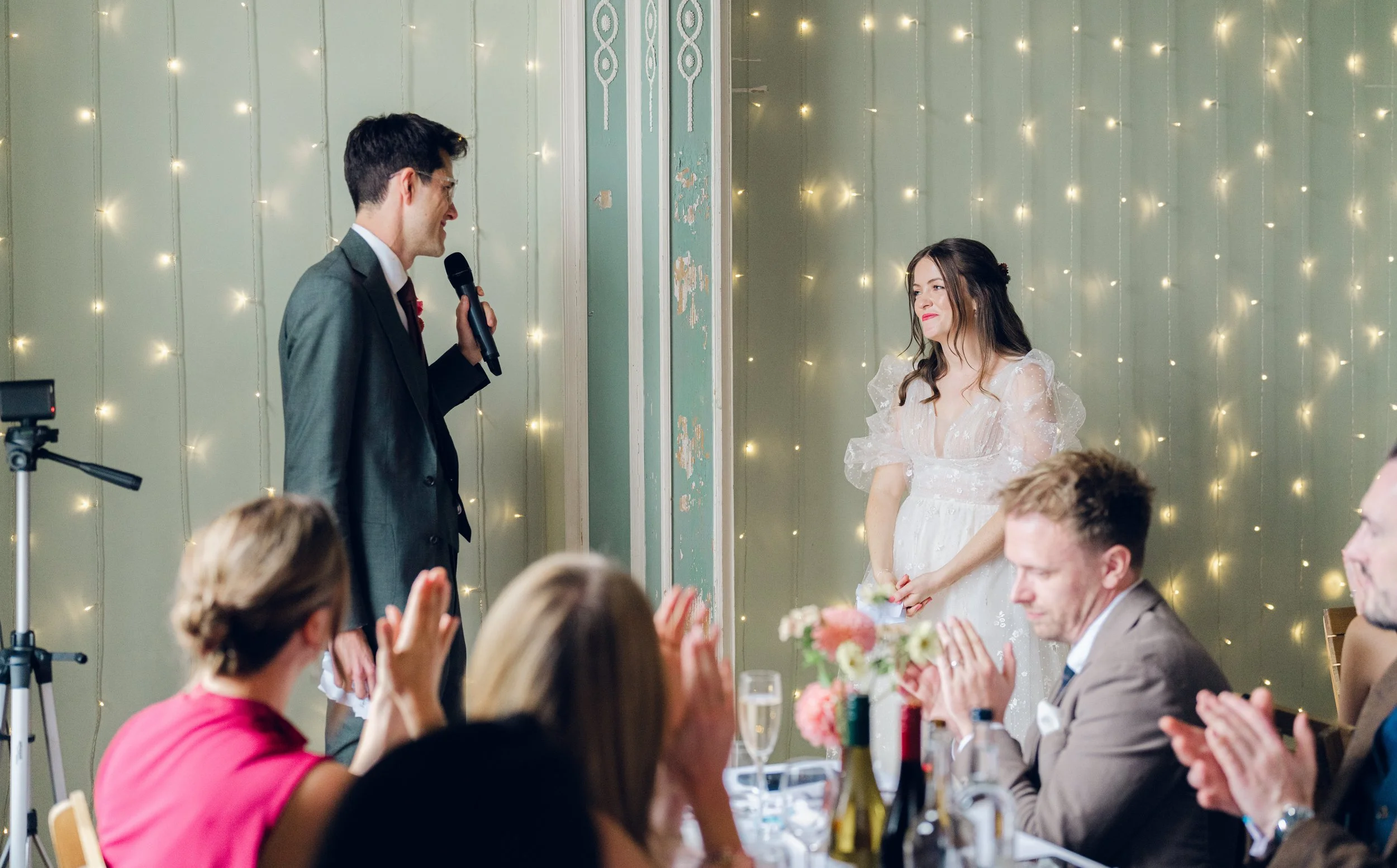 A man in a gray suit giving a speech at a wedding reception, with a woman in a white dress standing nearby and smiling. Guests seated at tables clapping and watching, decorated with flowers and fairy lights.