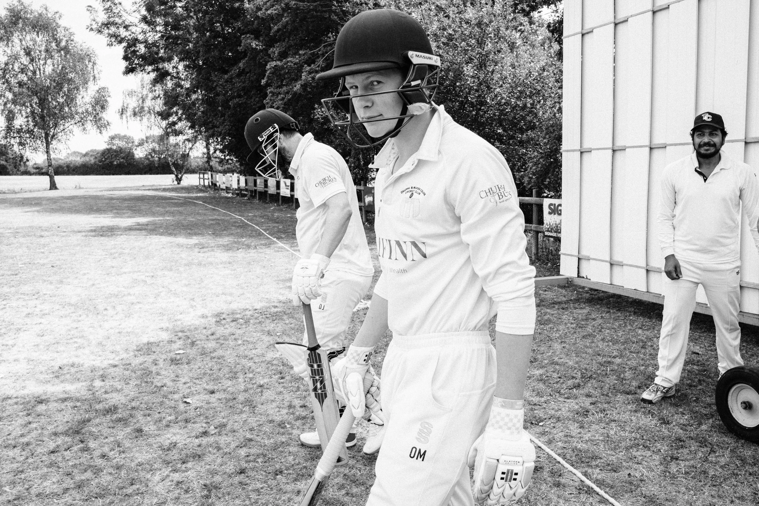 Cricket players standing on the cricket field, wearing helmets and cricket gear, with one player holding a cricket bat and another with a cricket ball, near a boundary line with trees in the background.