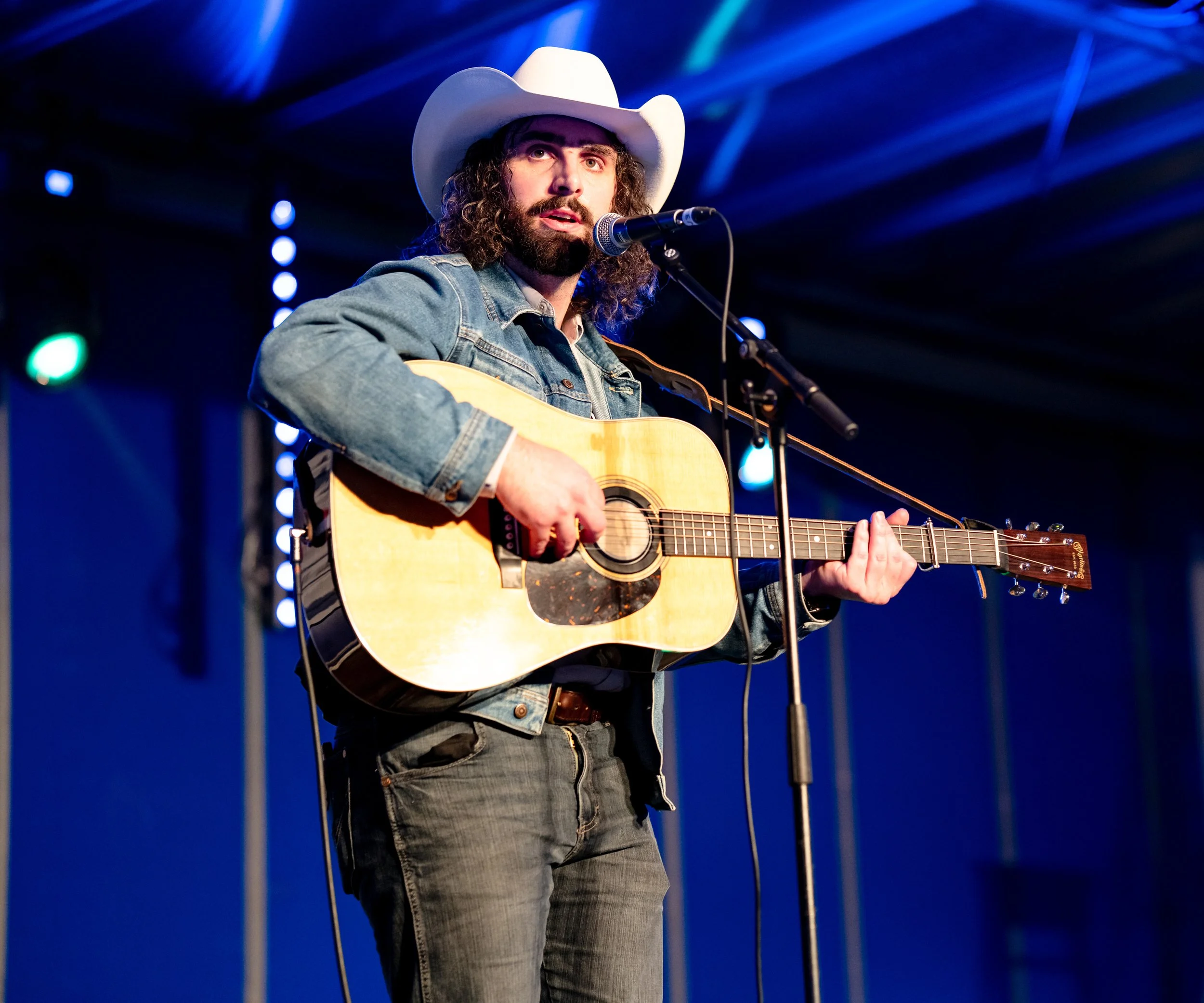 A man with long curly hair and a beard, wearing a white cowboy hat and denim jacket, playing an acoustic guitar and singing into a microphone on stage with blue stage lighting.