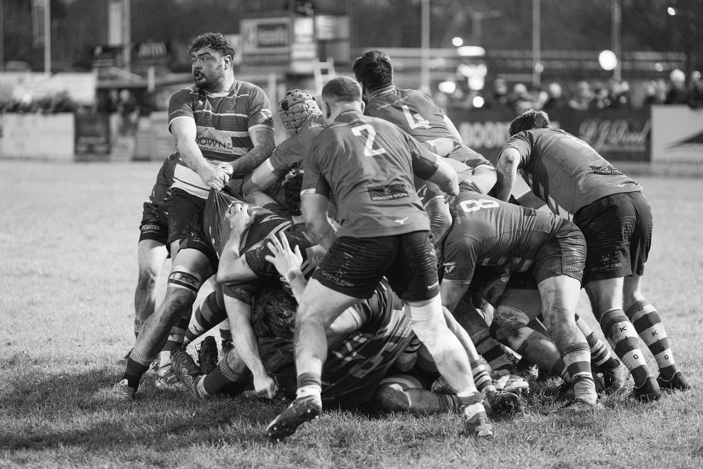 A black and white photograph of rugby players engaged in a scrum during a match on a grass field. The players are wearing sports uniforms with some dirt on their uniforms and legs.