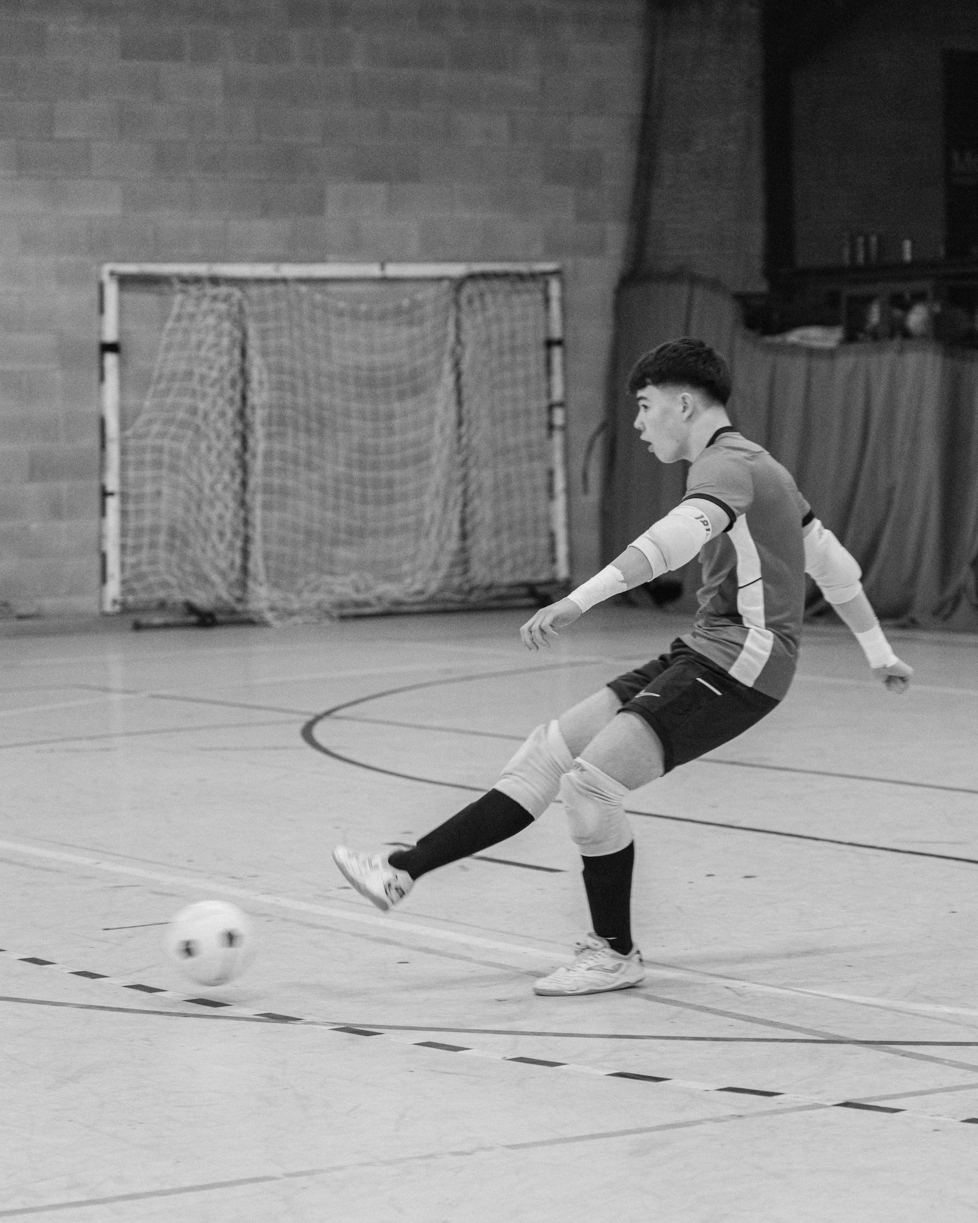 A young man in sportswear playing indoor soccer, kicking a ball on a court with a soccer goal in the background.