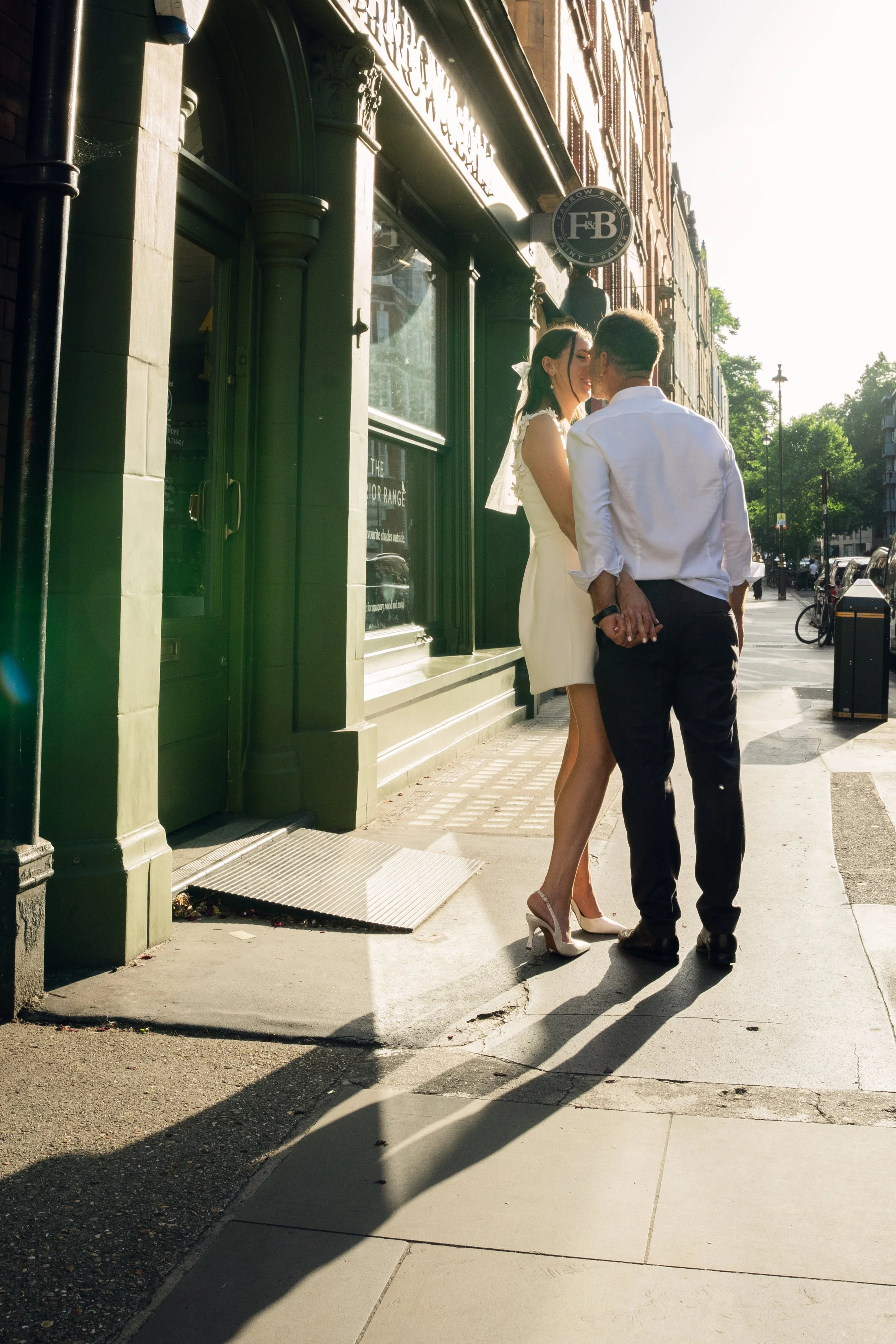 A couple standing on a city sidewalk, holding hands and leaning close to each other, with a building and a sign that reads 'F&B' in the background. The woman is wearing a white dress and high heels, and the man is in a white shirt and black pants. The scene is illuminated by sunlight, casting long shadows.
