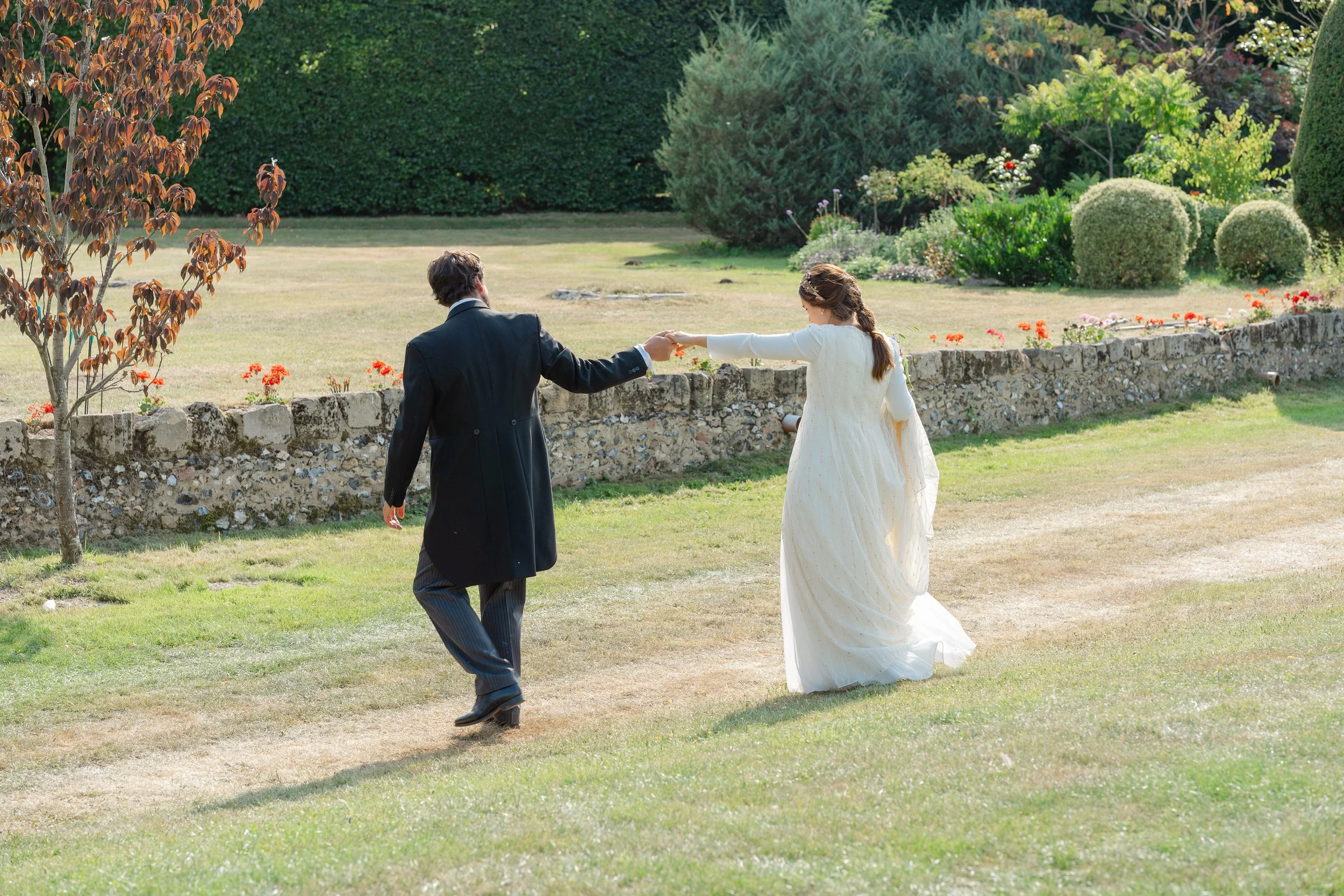 A bride and groom holding hands and walking together in a garden. The bride is wearing a white wedding dress and the groom is in a dark suit. The garden has a stone wall, green bushes, and colorful flowers.