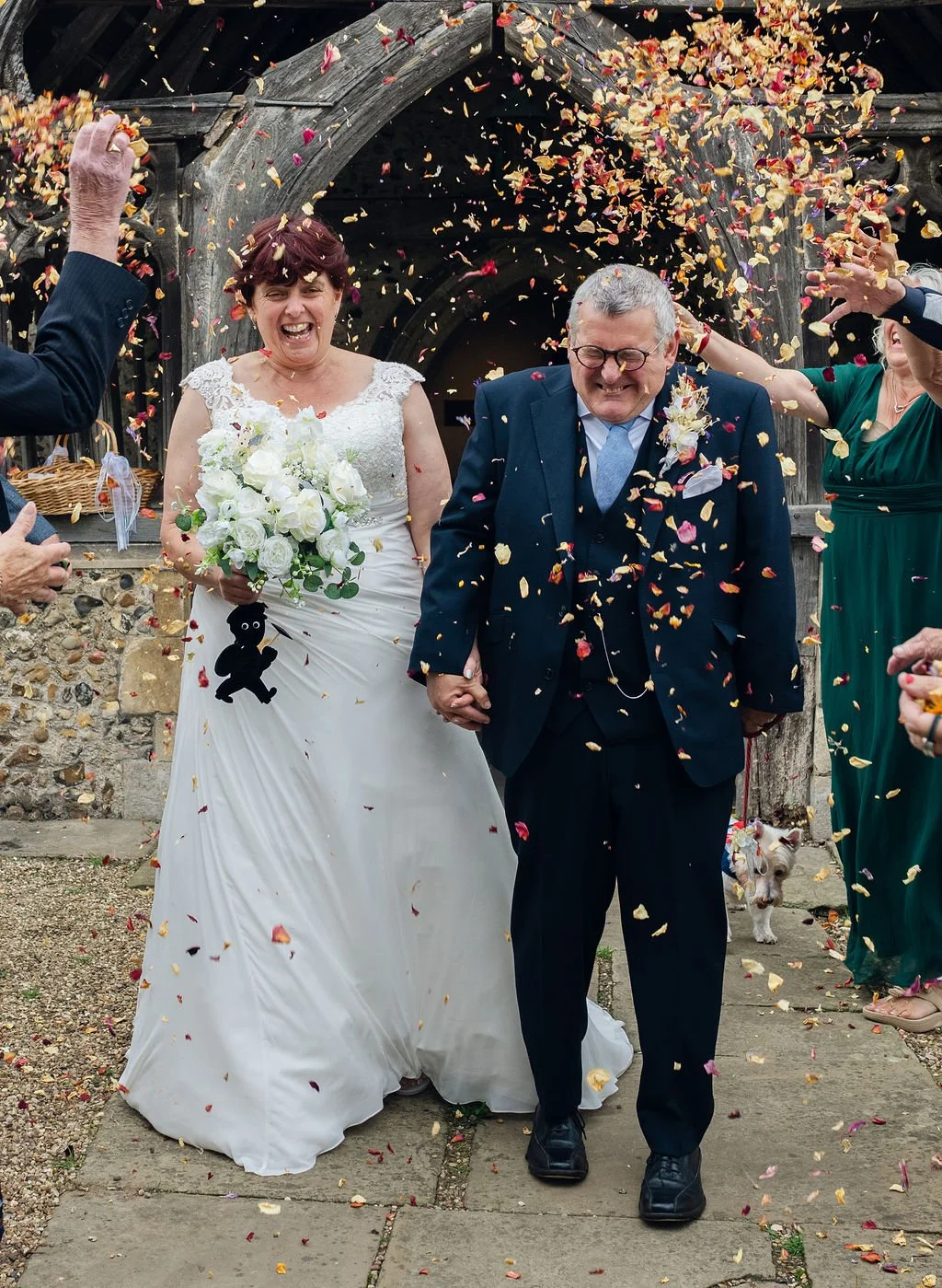 A bride and groom celebrating their wedding day, walking hand in hand while surrounded by guests throwing confetti. The bride is smiling and holding a bouquet of white roses, wearing a white dress with lace details. The groom is wearing a navy blue s