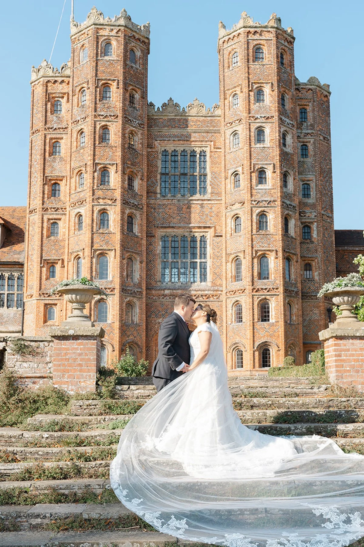 A bride and groom kissing on stone stairs outside a large brick castle with tall towers and many windows. The bride wears a white wedding gown with a long, flowing train. The groom is dressed in a dark suit.