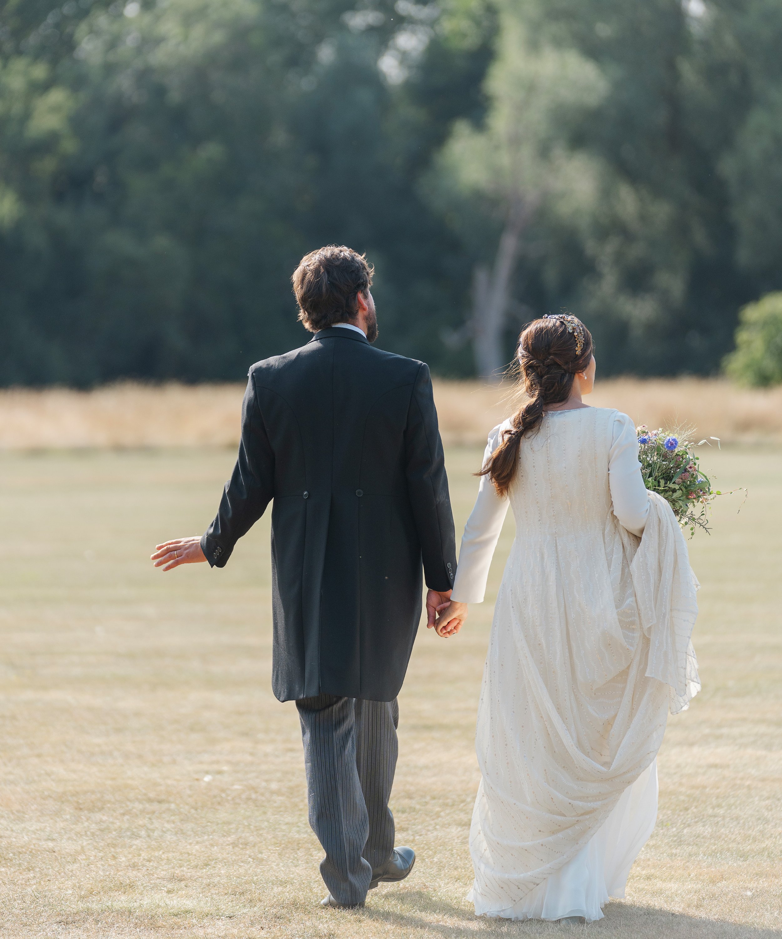 A bride and groom walking hand in hand across a field, holding a bouquet, with trees in the background.
