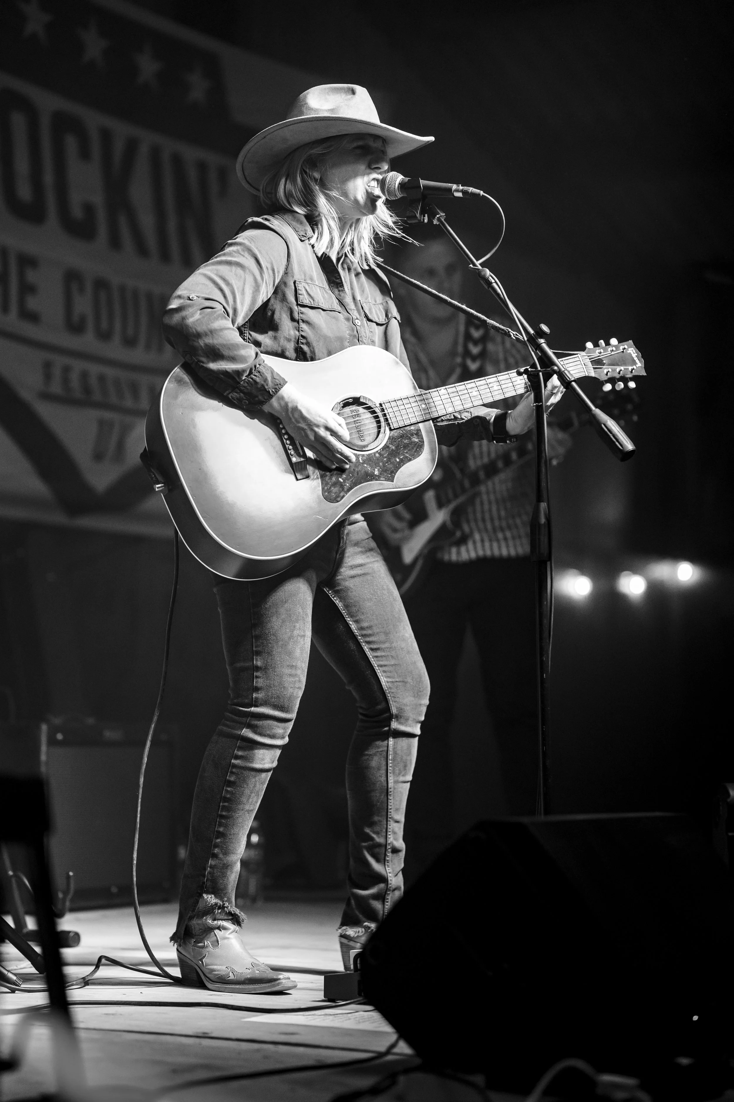 Female singer-songwriter performing on stage with an acoustic guitar, wearing a cowboy hat, denim jacket, ripped jeans, and high-heeled shoes, with a microphone in front of her.