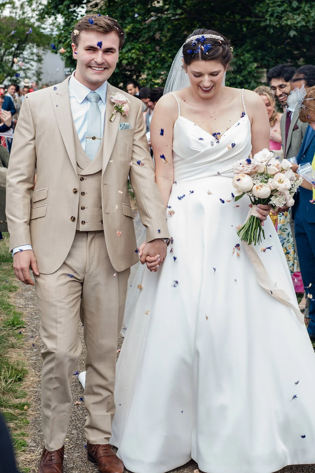 A newlywed couple holding hands during their wedding celebration outdoors, surrounded by guests, with confetti falling around them.