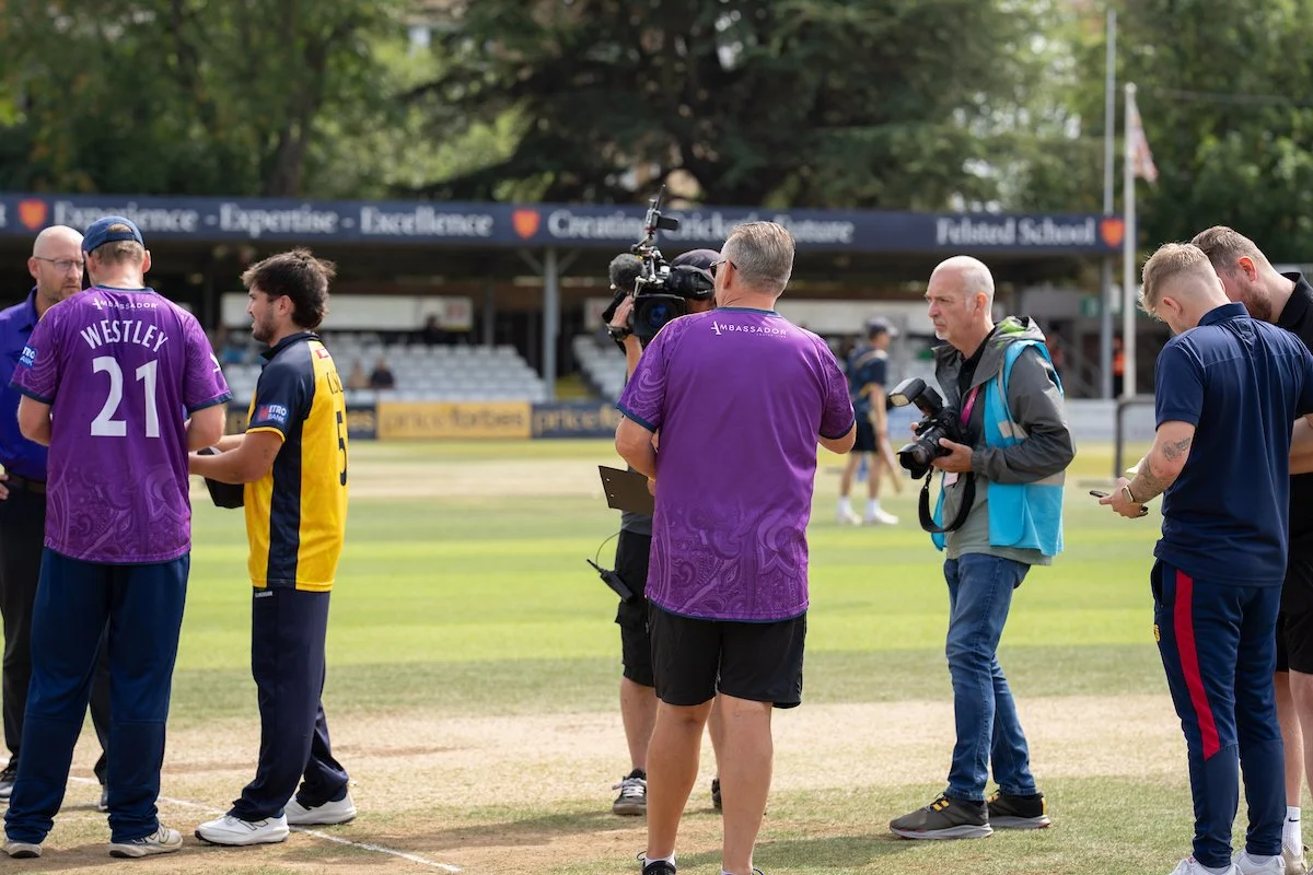 A group of people, including photographers and a man in a purple sports jersey, standing on a cricket field near the pitch, with a covered seating area and trees in the background.