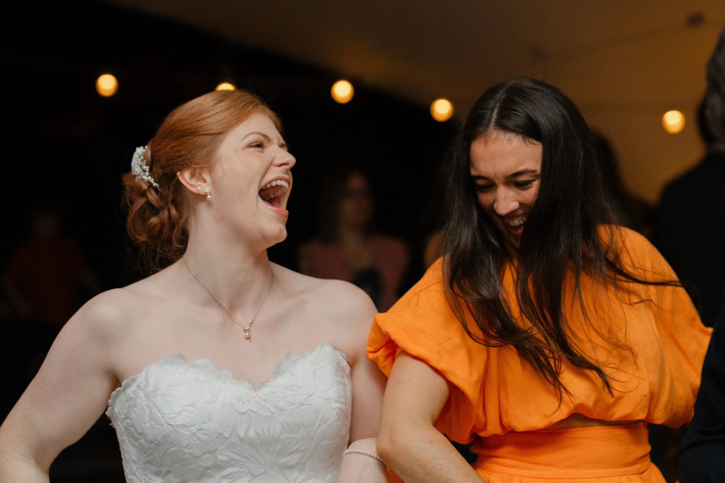 Two women laughing and having fun at a celebration or party, with one in a white strapless dress and the other in an orange dress, in a dimly lit indoor setting.