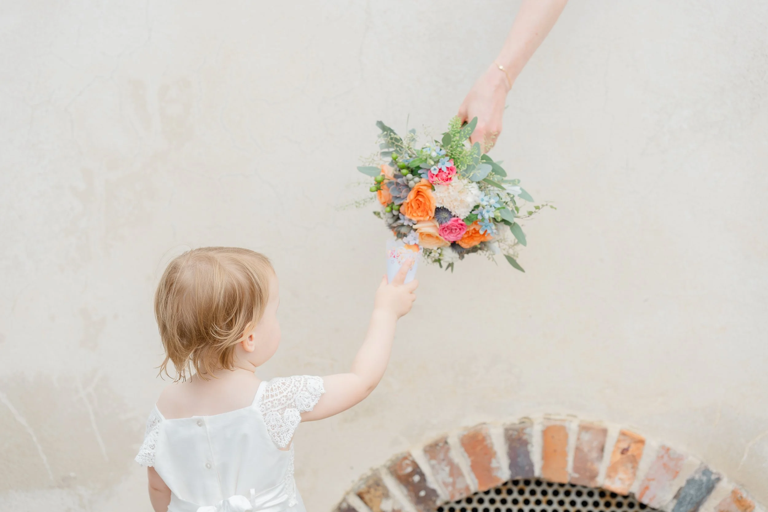 A young girl giving a bouquet of flowers to an adult, against a plain beige wall with a brick and metal vent near the bottom.