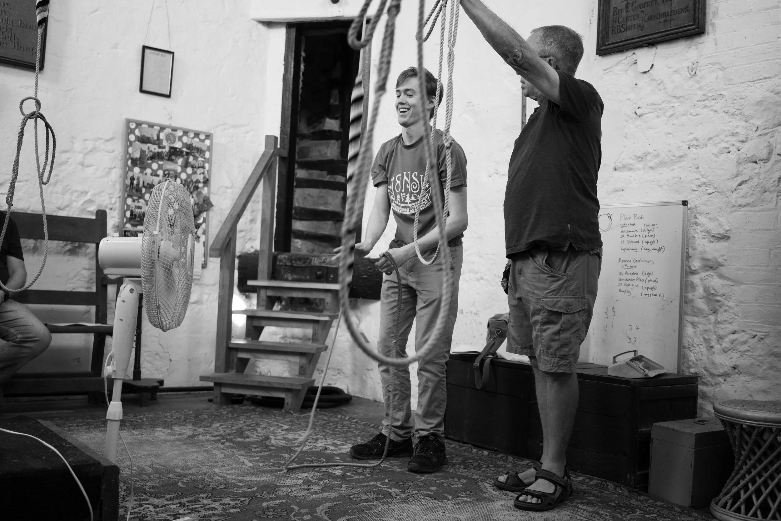 Two young men are standing indoors in front of a white brick wall. They are laughing and holding ropes, possibly participating in a game or activity. There is a staircase and framed pictures or signs on the wall behind them, along with a whiteboard on the right side with handwritten notes.