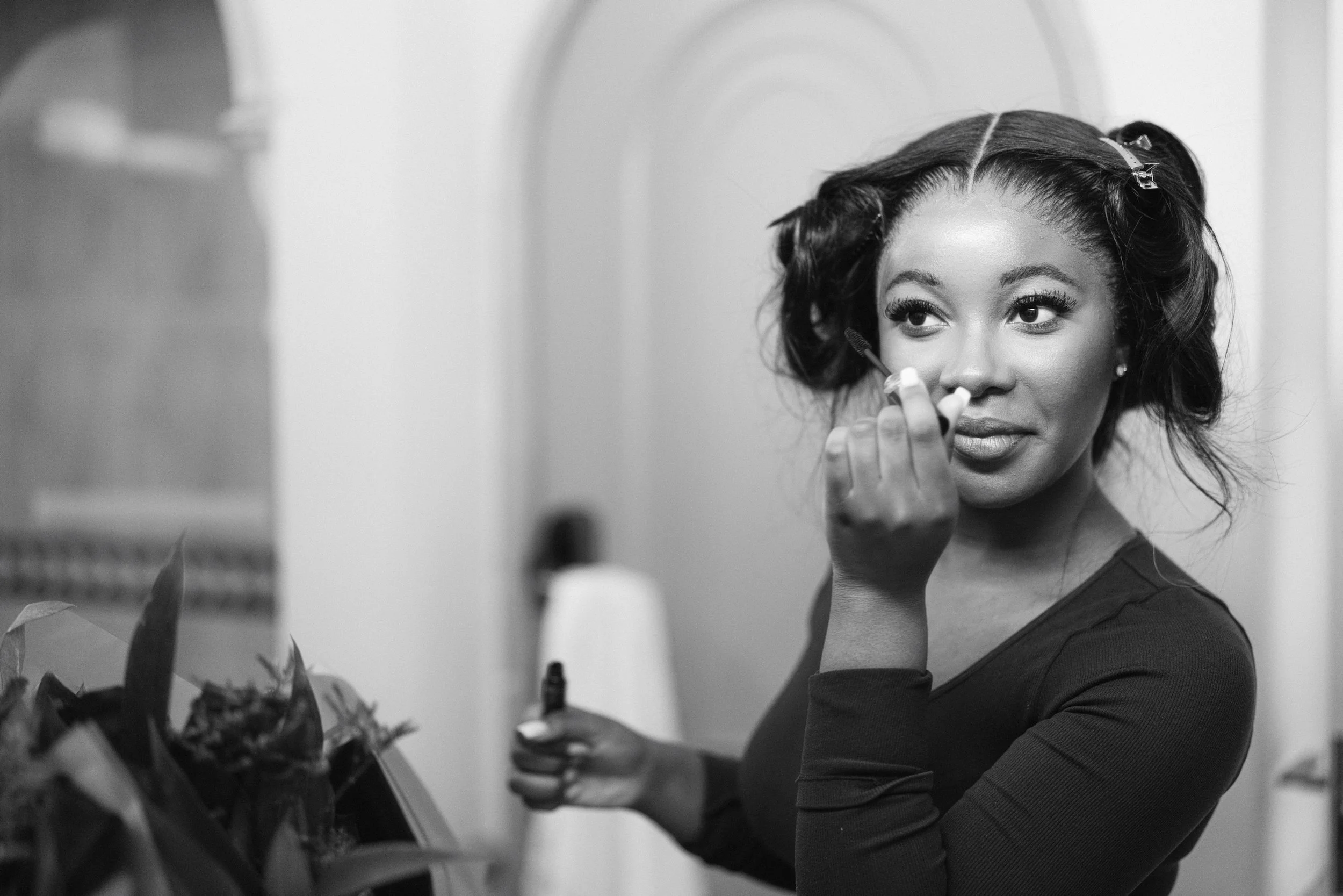 A woman with styled hair is applying makeup near a mirror in a bathroom or dressing room.