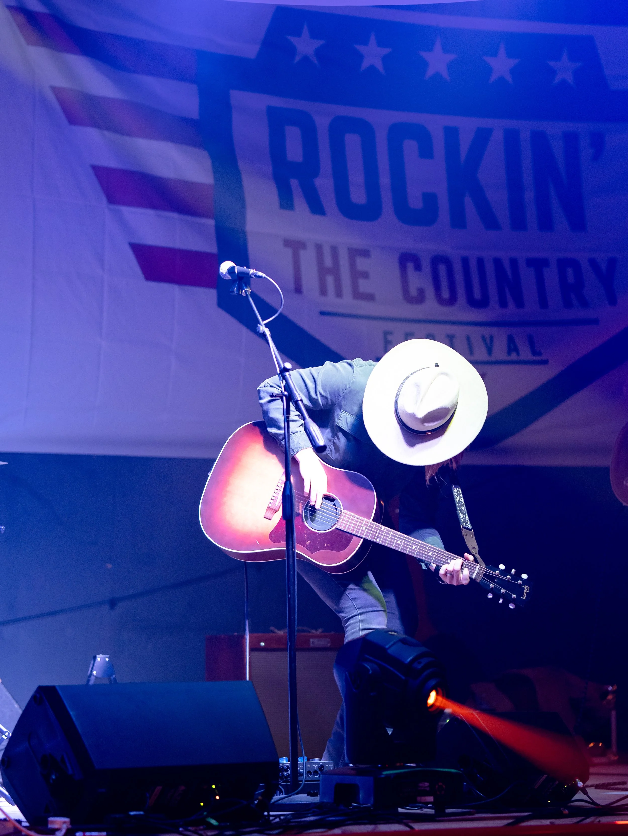 A musician wearing a large white hat and a dark jacket is playing an acoustic guitar on stage at the Rockin the County Festival, with a microphone in front of them. The background features a large banner displaying the festival's name.