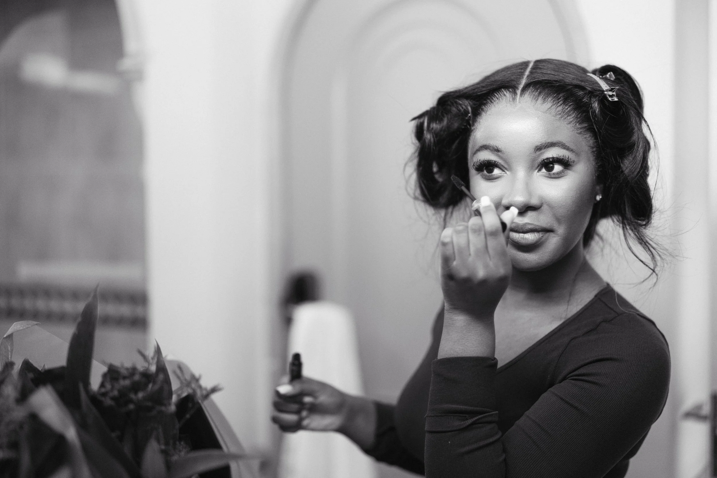 Young woman applying makeup in front of a mirror, holding a makeup product in one hand and using a brush with the other, with hair pinned back.