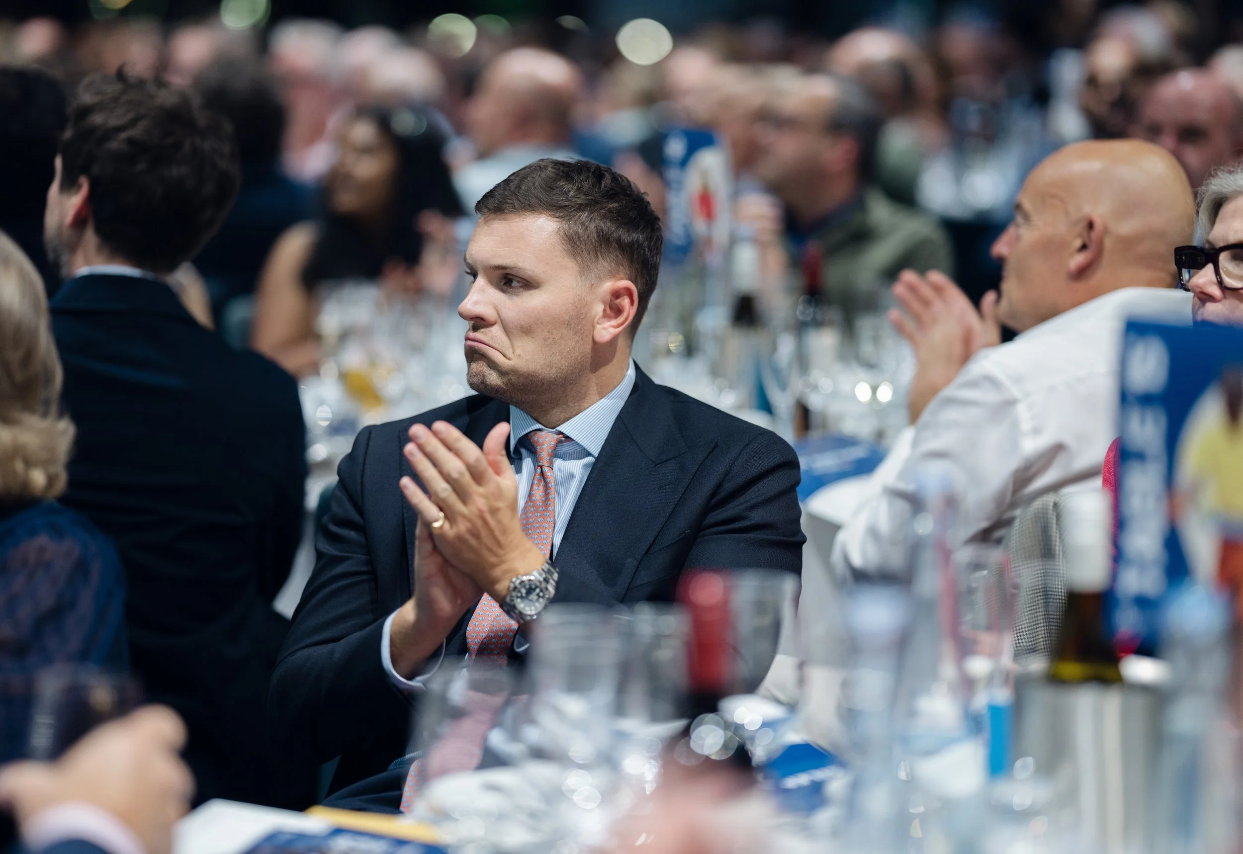 A man in a suit and tie sitting at a conference table clapping with a serious expression, amidst a large crowd of people at a formal event.