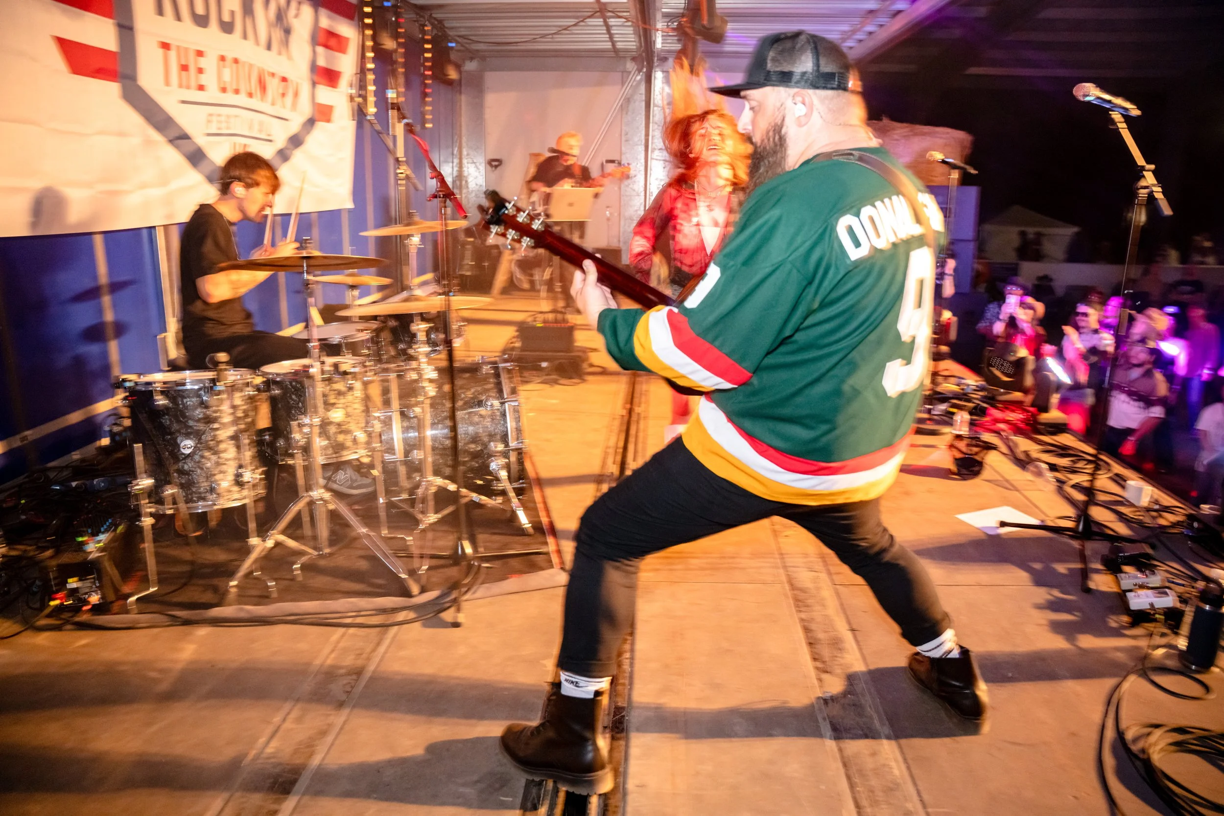 Musicians performing on stage at a live event, featuring a drummer playing drums and a guitarist playing guitar, with an audience watching in the background.