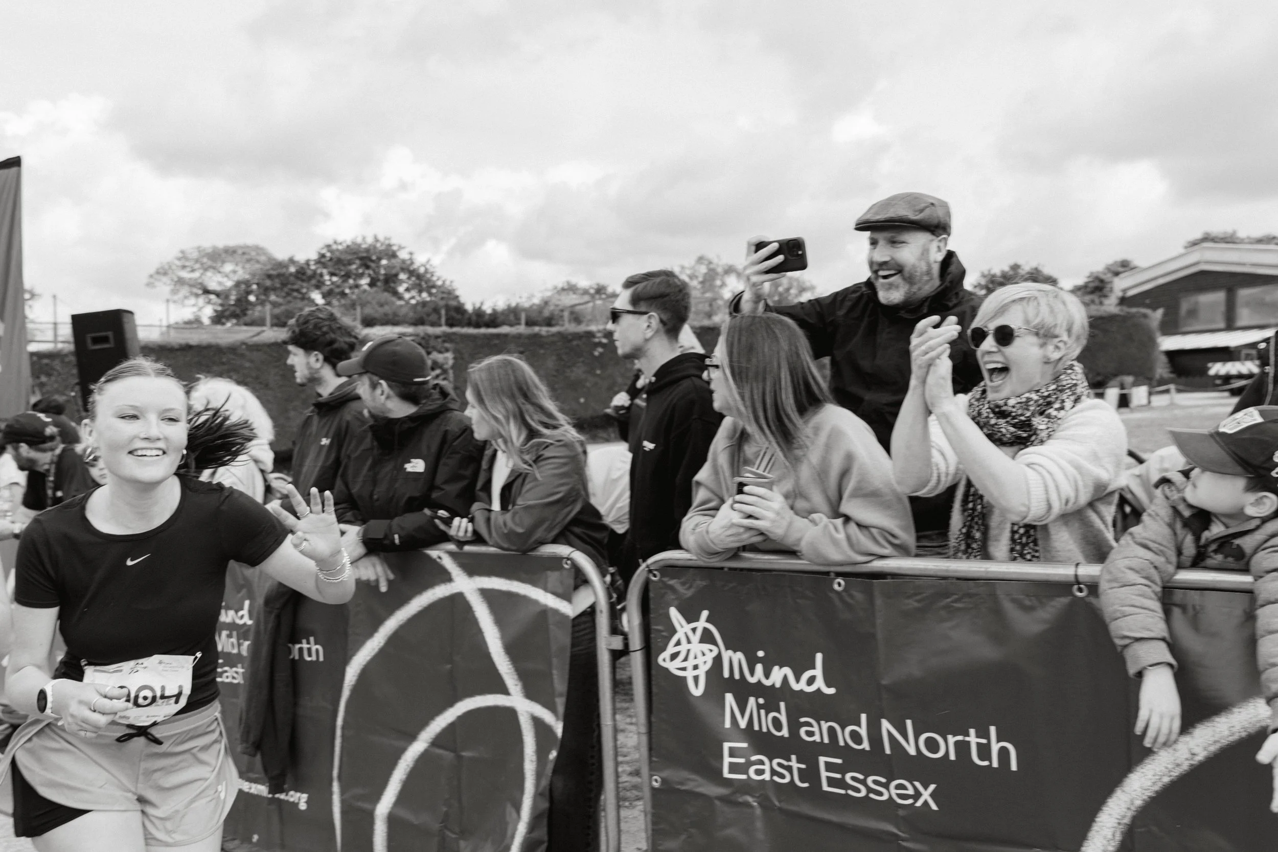 People participating in an outdoor event or race, with some smiling and looking cheerful, behind a barrier with a banner that reads 'Mind Middle and North East Essex.'