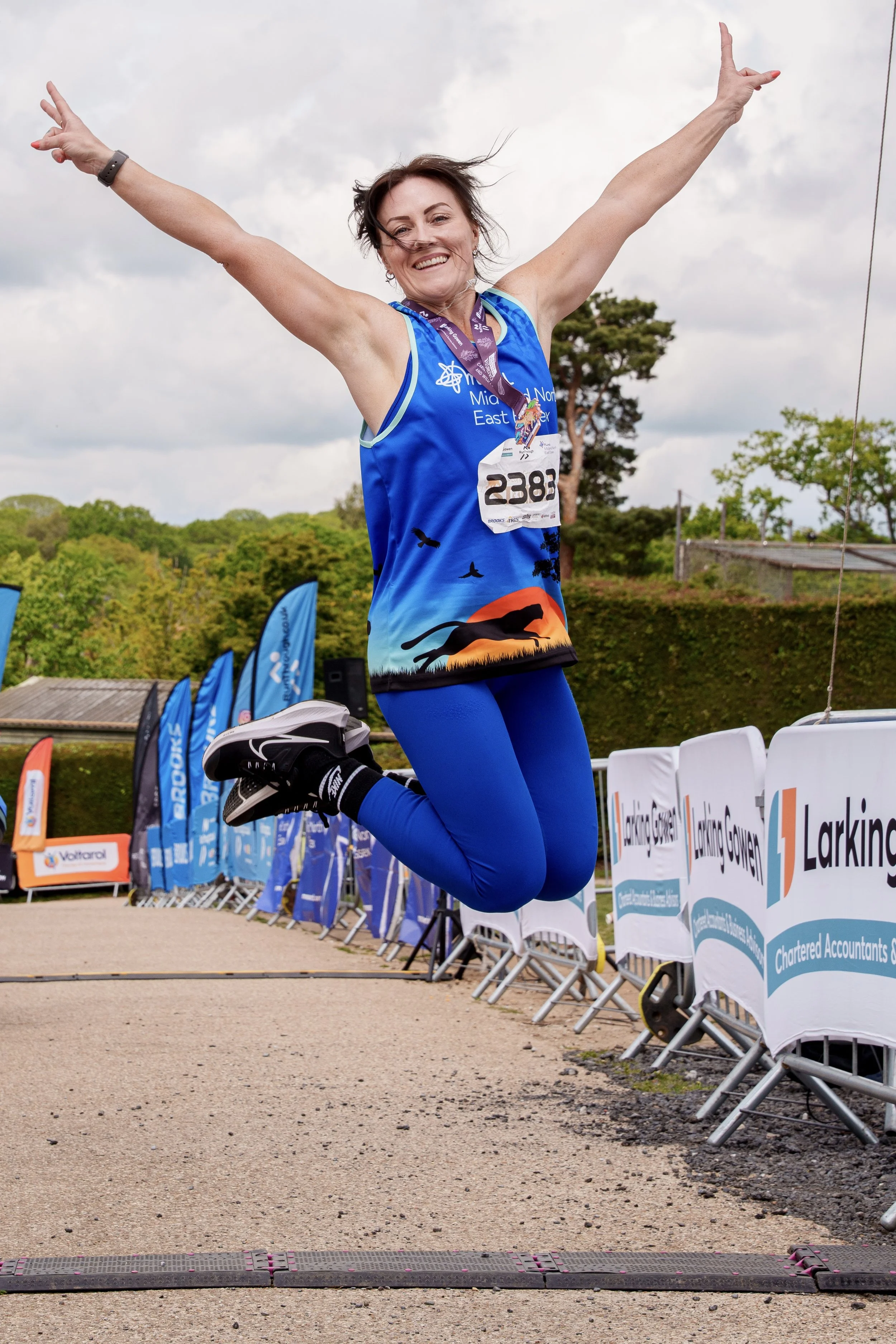 A woman wearing a blue running tank top and blue leggings jumping over a finish line at a race, smiling with her arms raised in victory. There are balloons and banners with sponsor logos in the background.