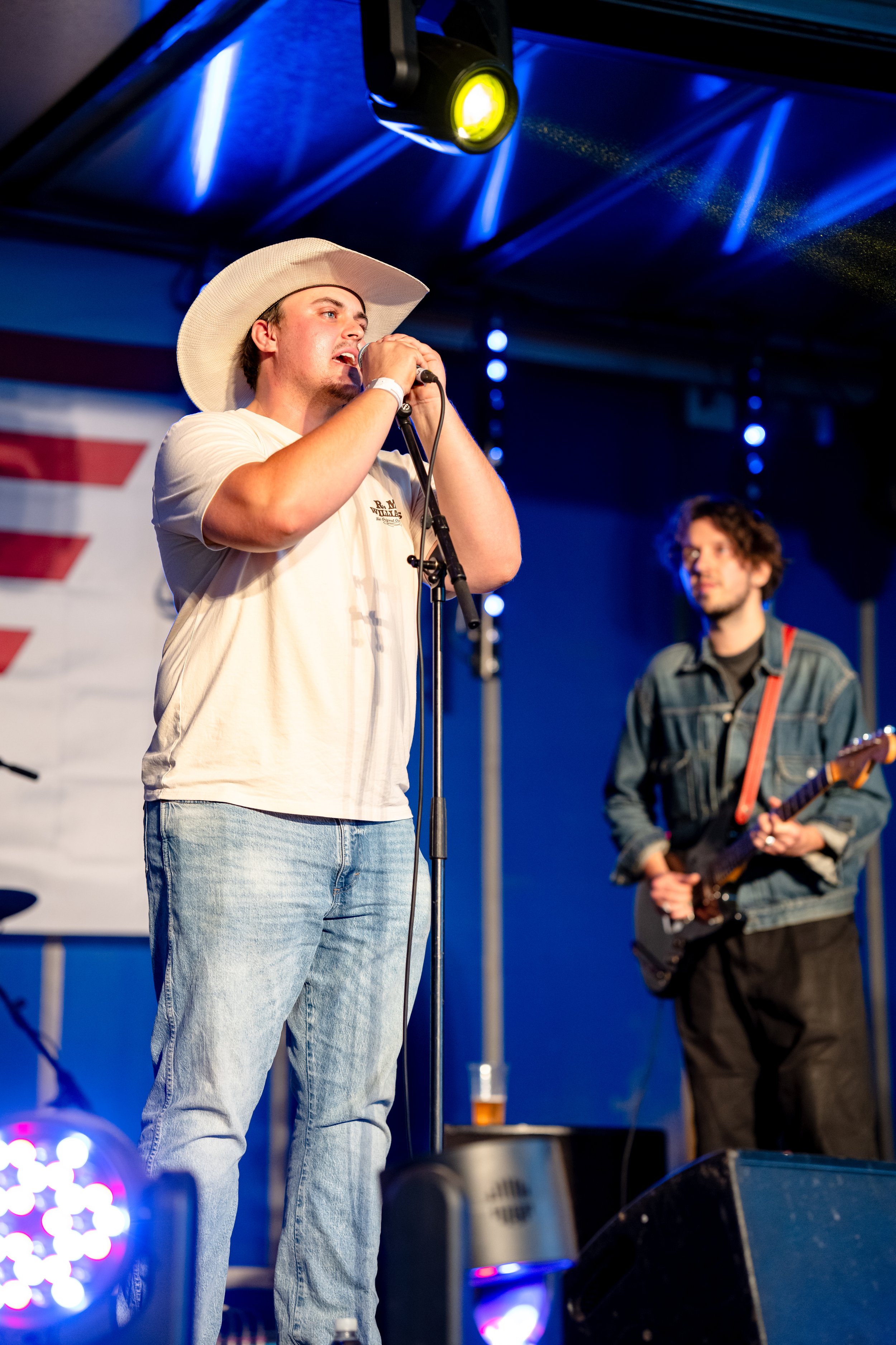 A young man wearing a cowboy hat singing into a microphone on stage, with another person in the background playing a guitar.