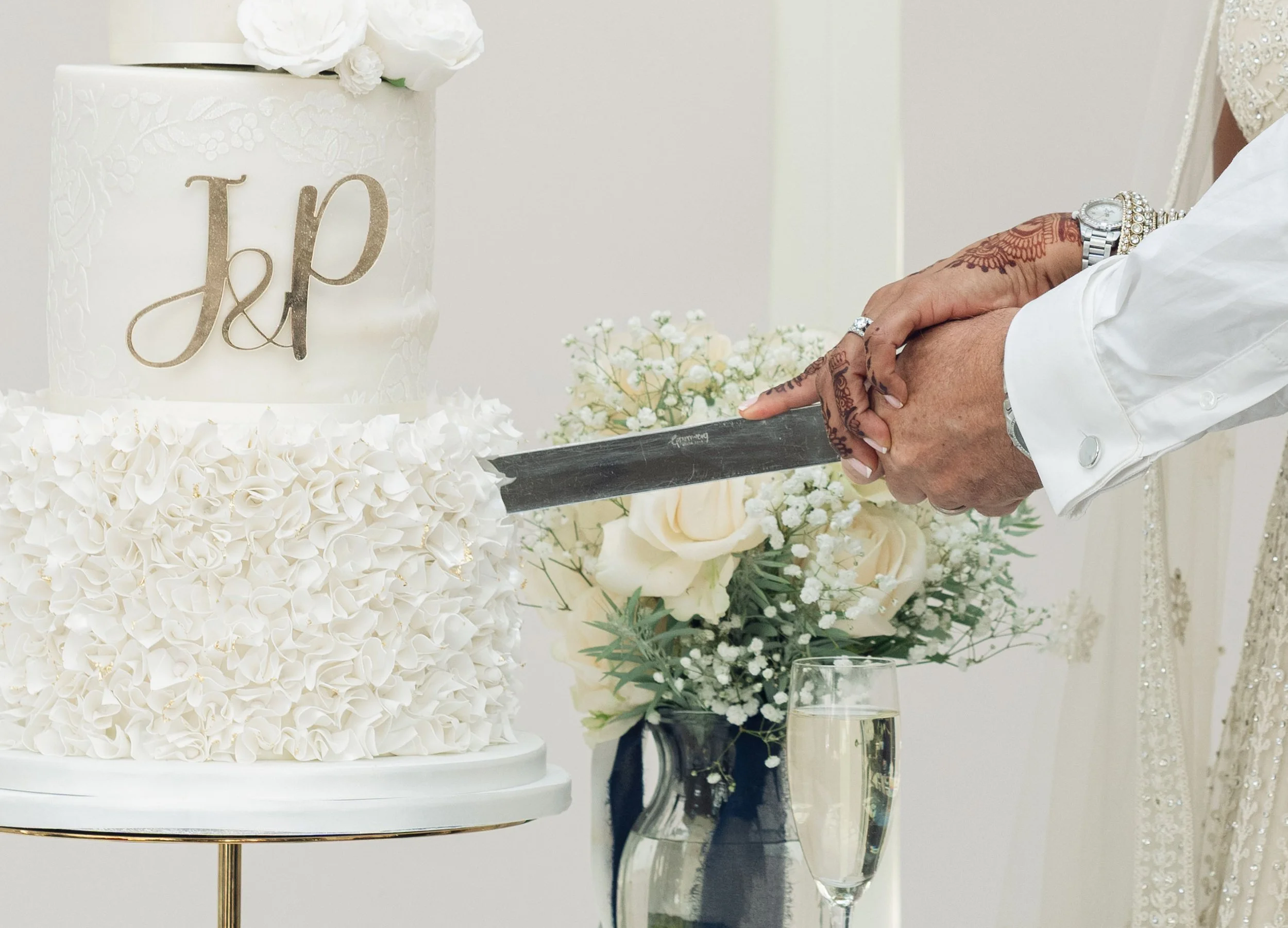 Person cutting a white wedding cake with a knife, decorated with floral patterns and the initials J&P. The cake is on a stand, surrounded by floral arrangements and a glass of champagne.