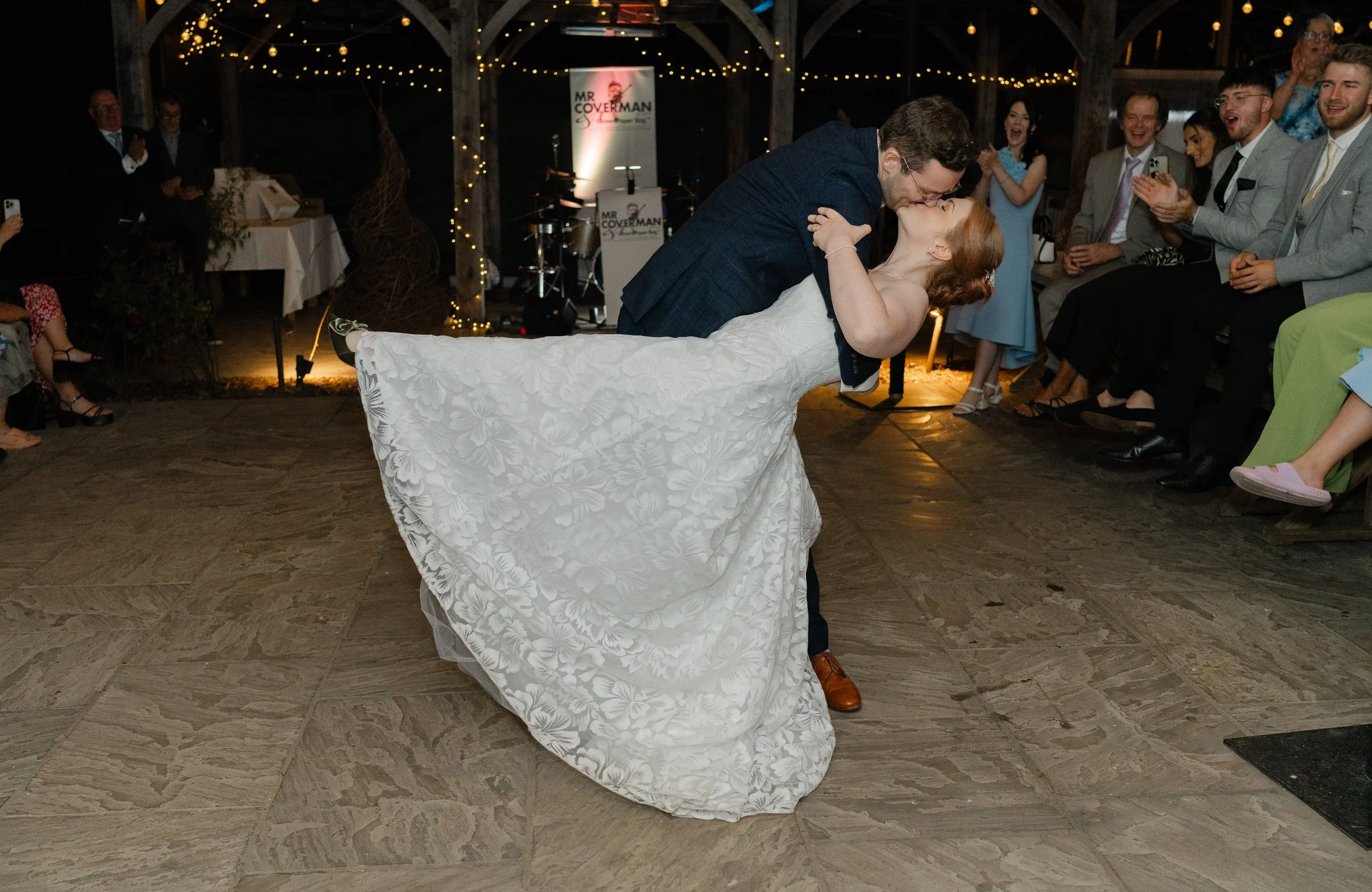 A groom dips and kisses his bride during their wedding dance, surrounded by seated guests in a decorated venue with string lights.
