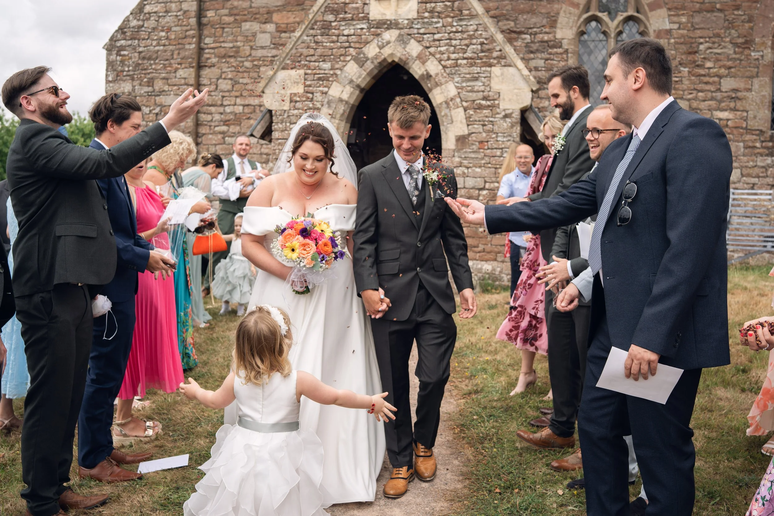 A bride and groom walking hand in hand through a crowd of guests outside a stone church during their wedding celebration. The bride holds a colorful bouquet and is smiling, while the groom also smiles as they walk. Guests on both sides are cheering a