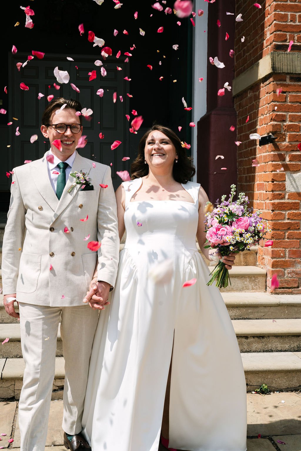 A newly married couple smiling and holding hands outside their wedding venue, surrounded by falling pink and white flower petals. The bride wears a white gown and holds a colorful bouquet, while the groom wears a cream-colored suit with a green tie.