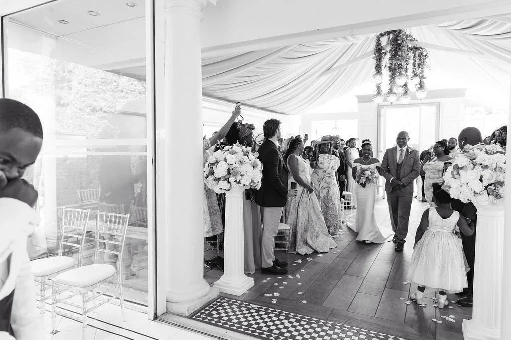 A black and white photo of a wedding ceremony with guests standing inside a decorated venue, some holding bouquets, and a woman in a wedding dress approaching the altar.