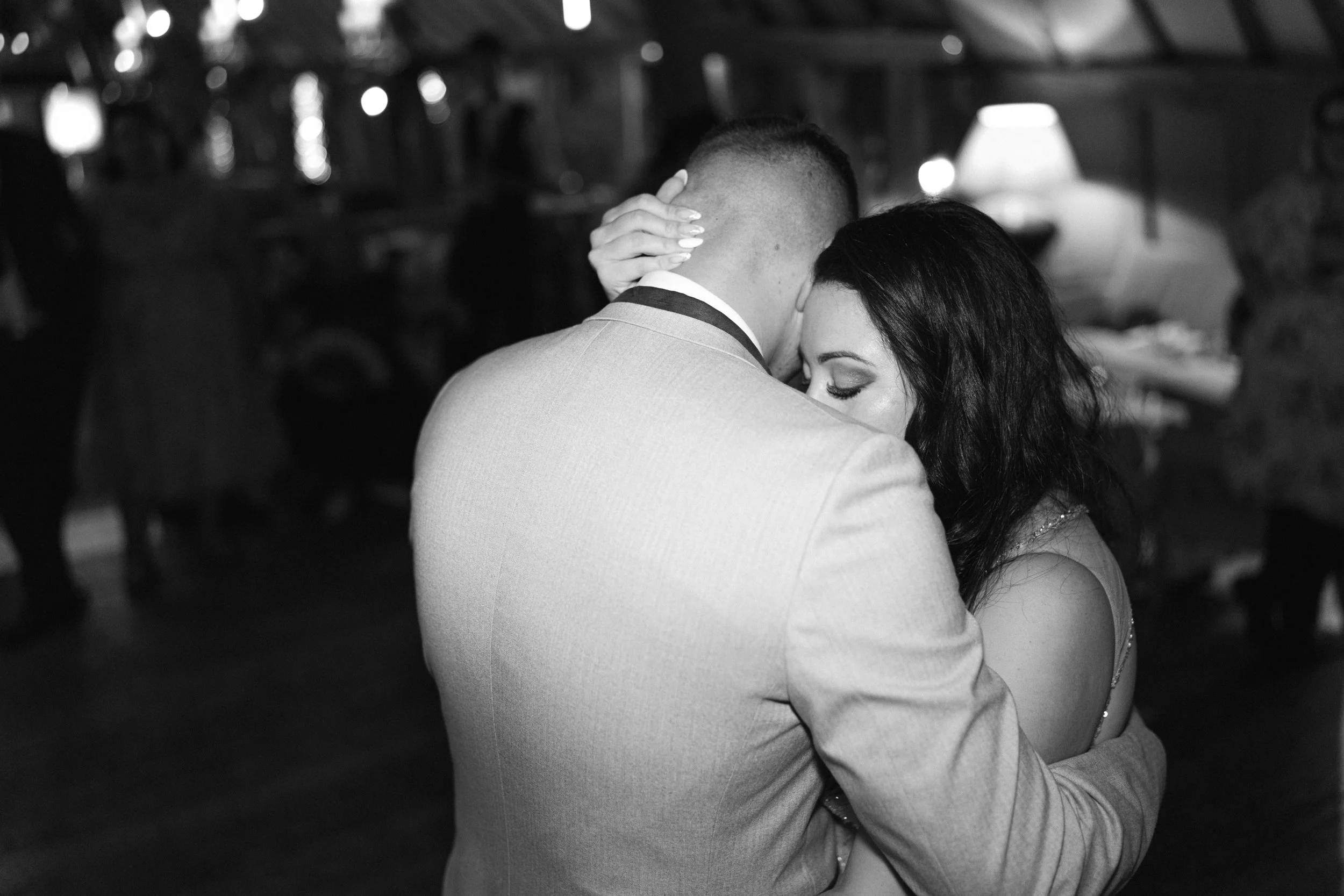 A black and white photo of a couple hugging closely, with their eyes closed, in a cozy indoor setting with blurred background.