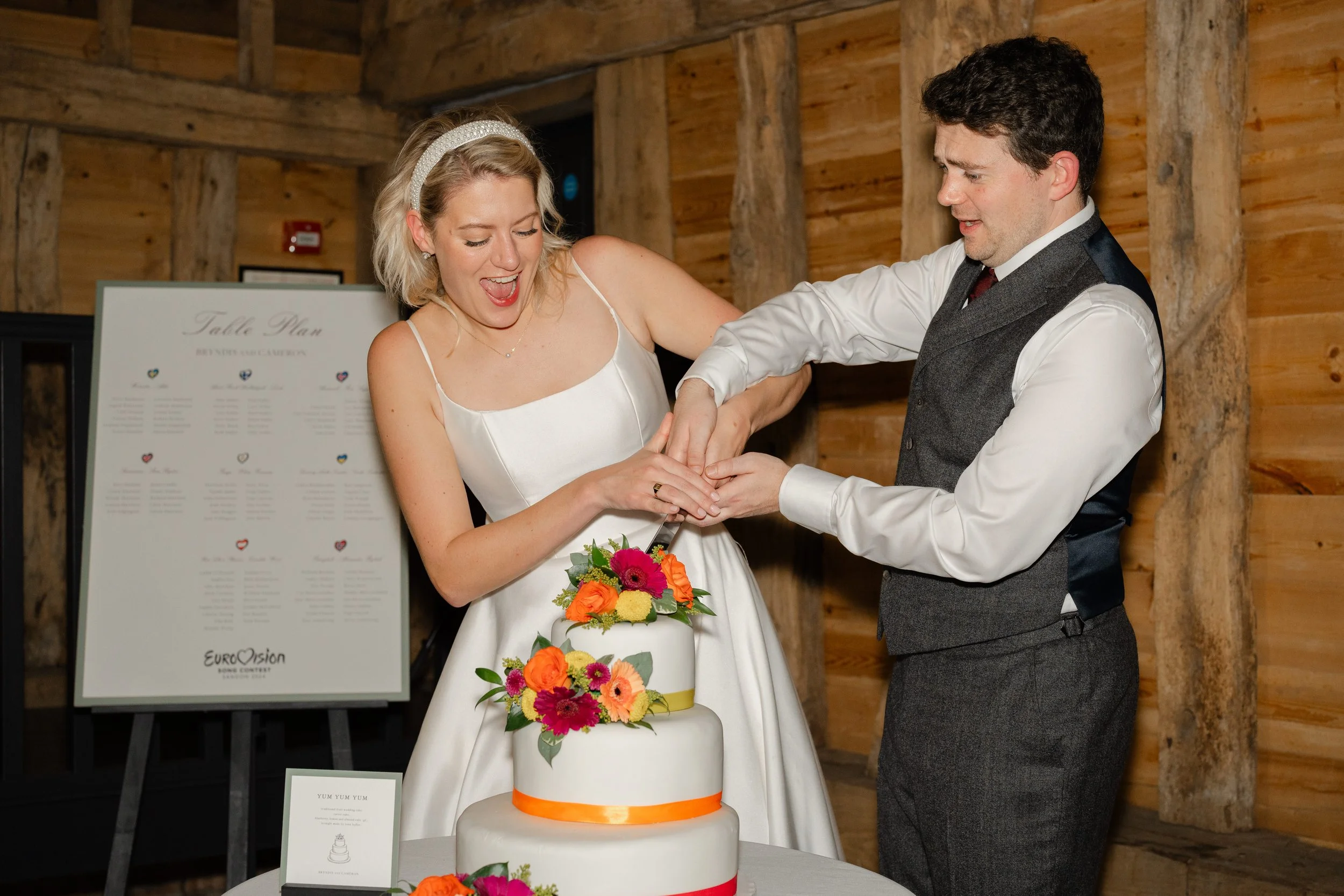 A bride and groom cut a wedding cake together in a wooden indoor setting, with the bride wearing a white satin dress and the groom in a white shirt and gray vest. The wedding cake is decorated with colorful flowers and orange ribbon accents, and they